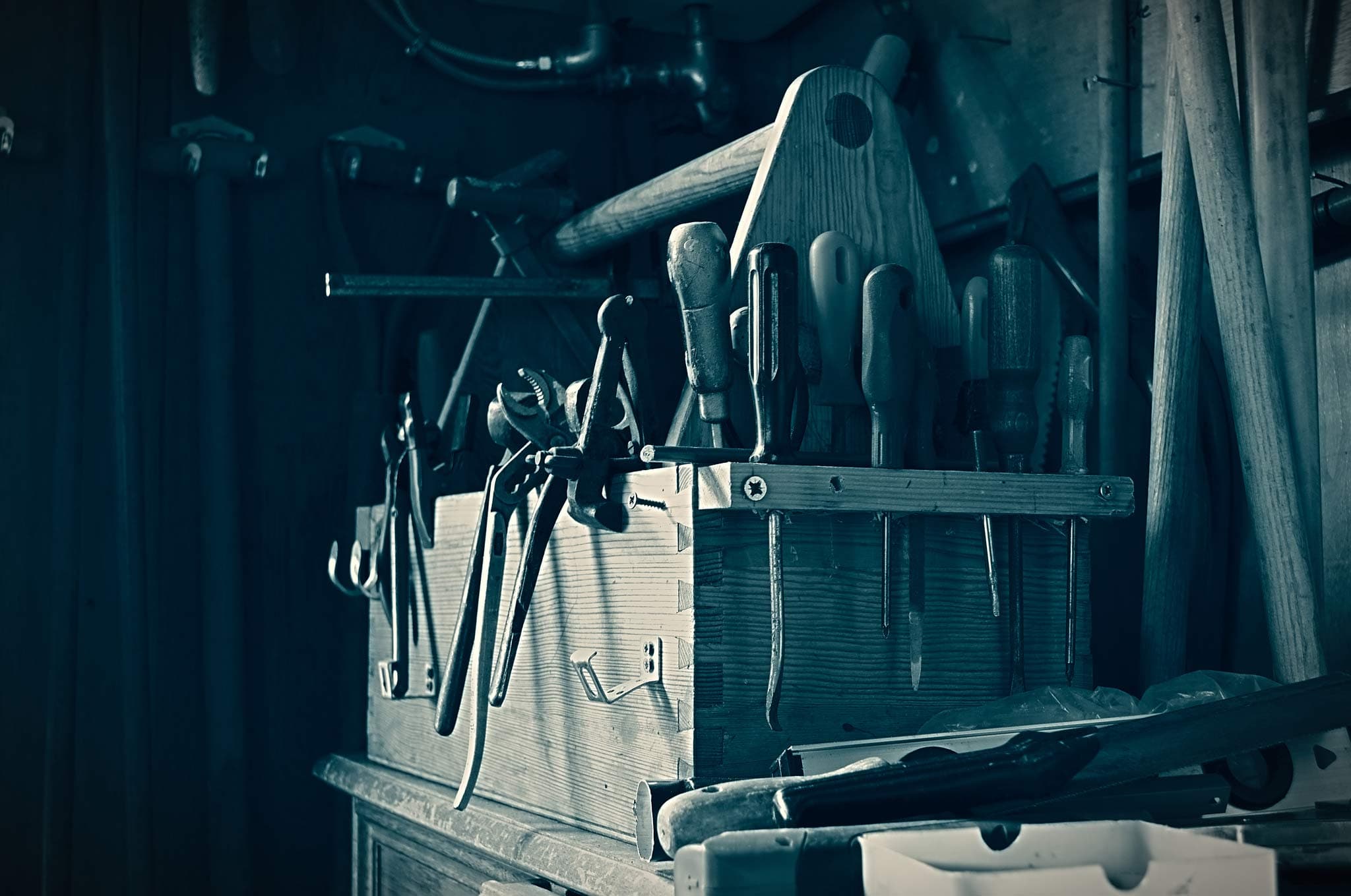 A wooden toolbox filled with various hand tools such as pliers and screwdrivers is on a workbench. The tools are arranged upright, and there are other tools like hammers and axes leaning against the wall in the dimly lit workshop.