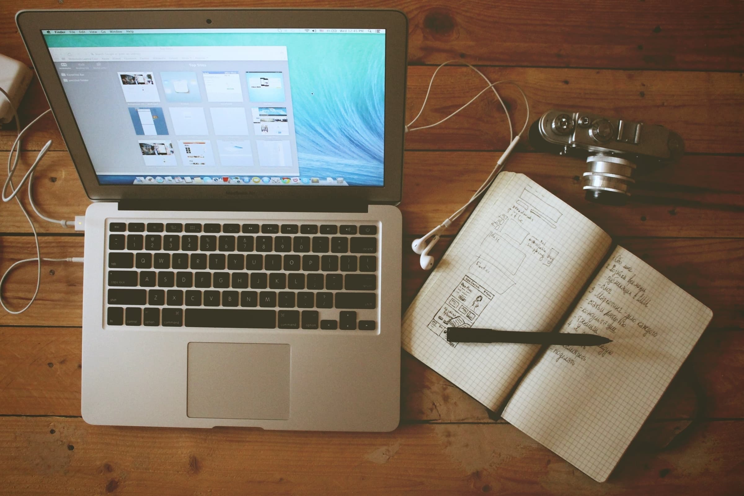 An open laptop on a wooden desk displaying a webpage layout. Nearby are white earphones, a vintage camera, and an open notebook with sketches and notes. A pen rests on the notebook. The scene suggests a creative workspace.