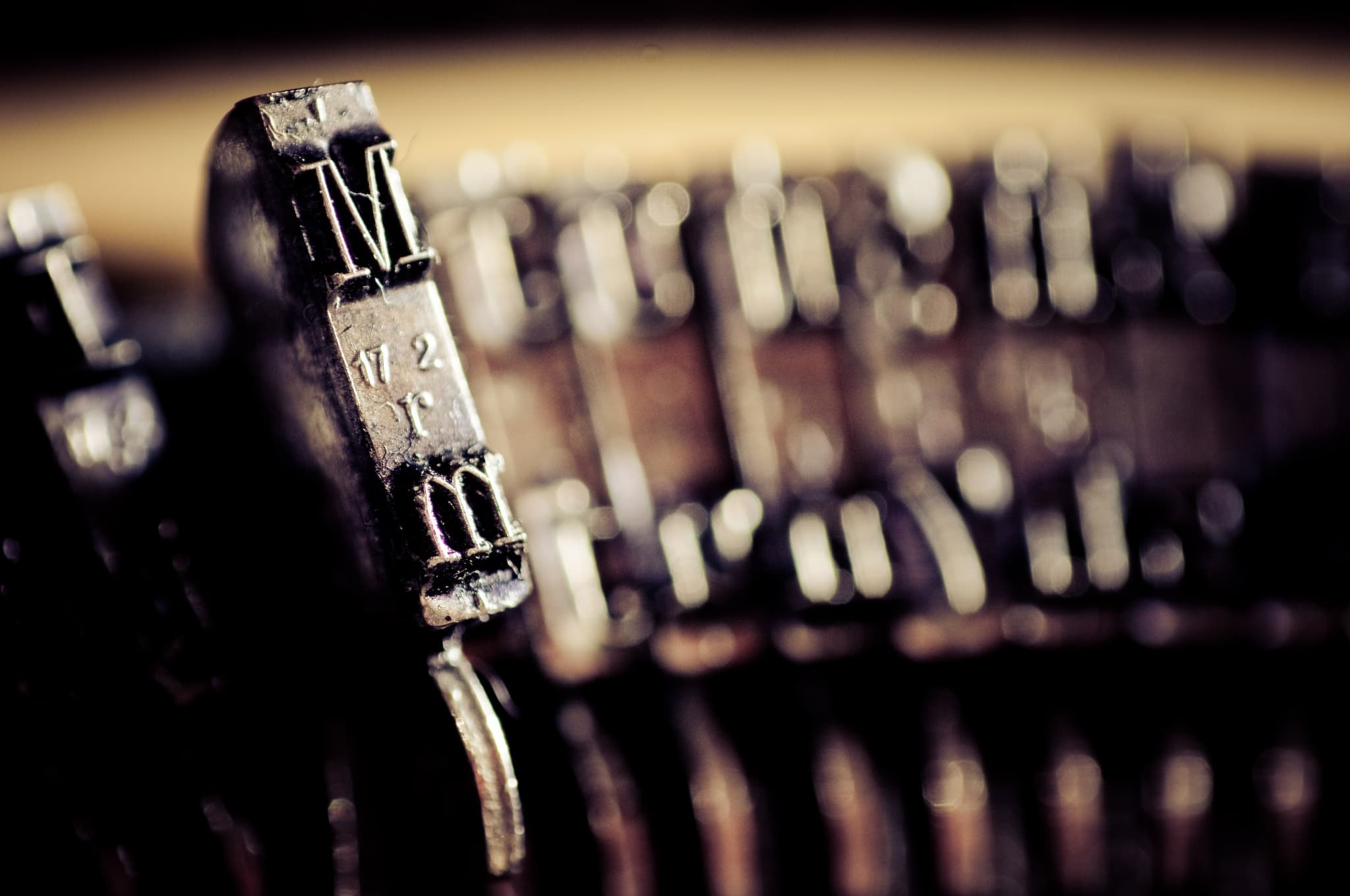 Close-up of vintage typewriter keys with the letter M in focus. The keys have a metallic, worn appearance, with the background keys blurred, giving a sense of depth and age.