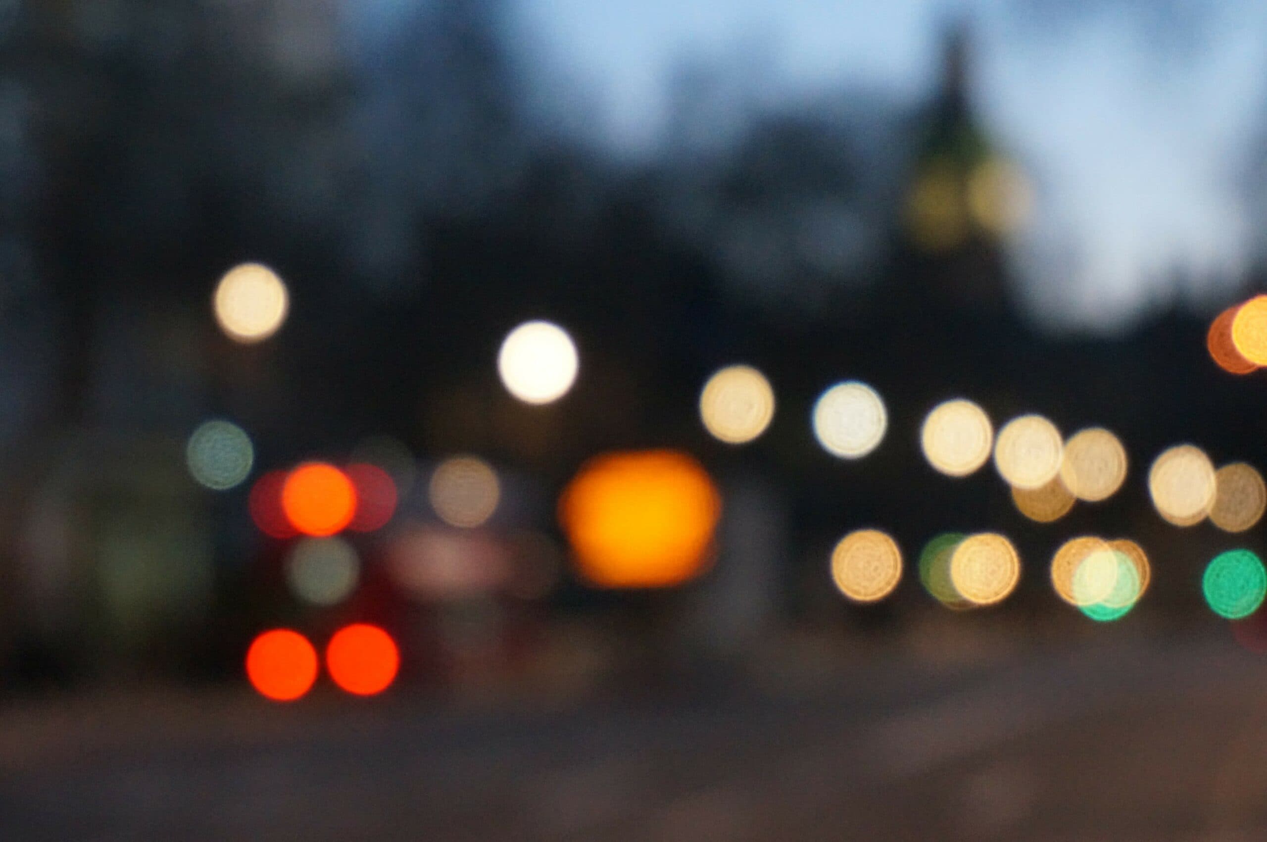 A blurred cityscape at dusk featuring circular bokeh lights in various colors. The scene includes indistinct shapes, possibly a road and a vehicle, set against a fading blue sky.