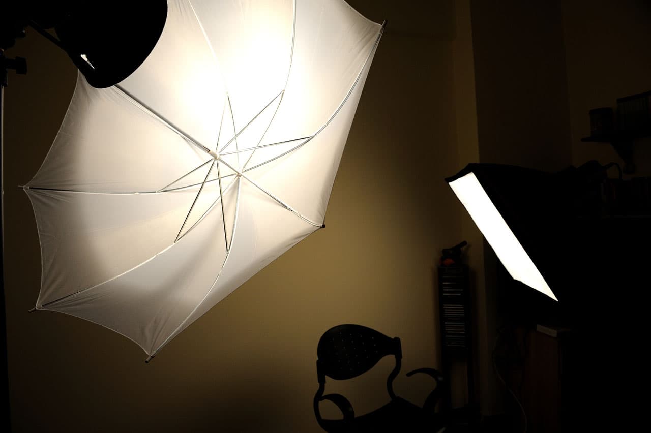 A photography studio setup with a white umbrella light on the left and a rectangular softbox on the right. A black chair is positioned near the center against a dimly lit background.