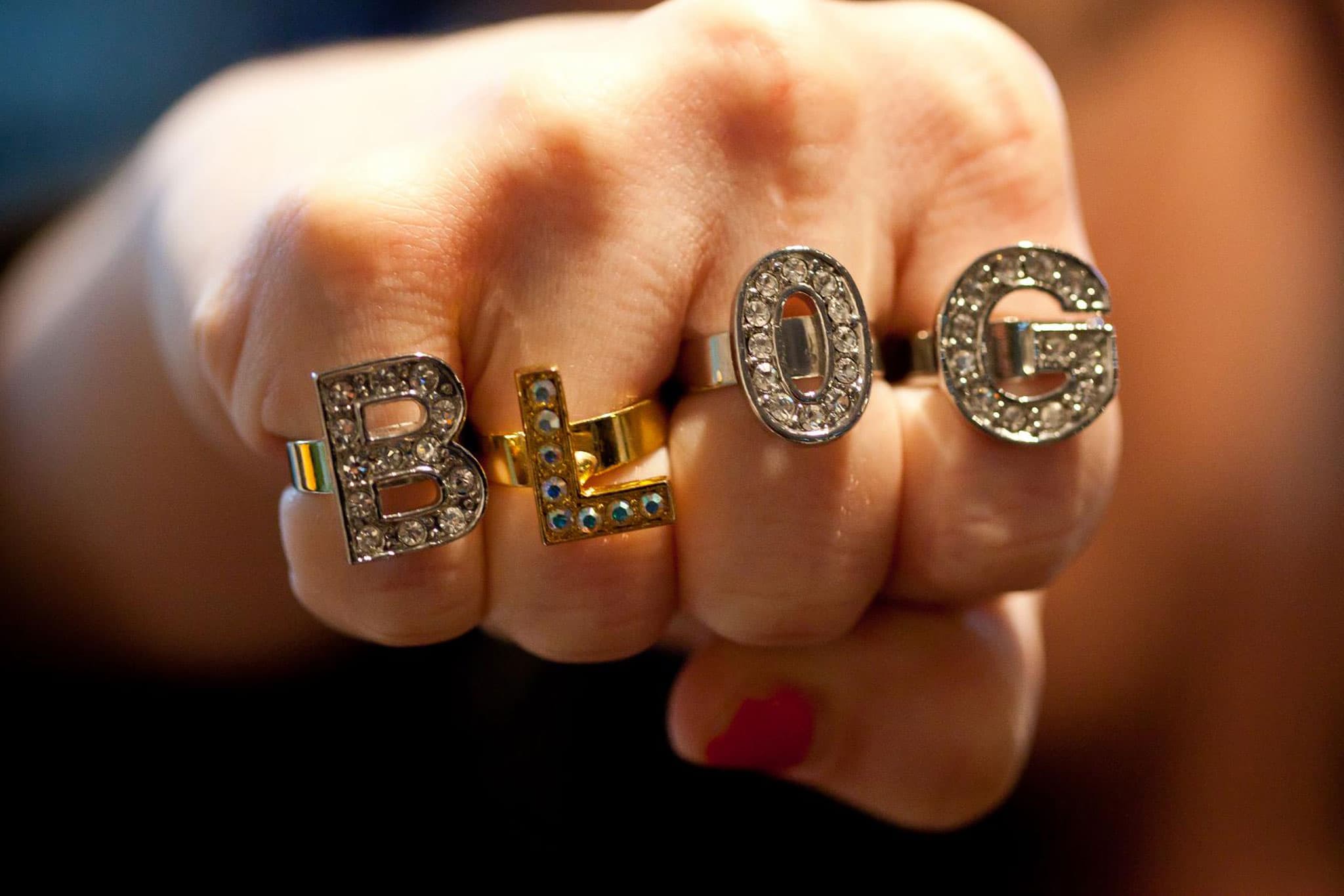 A close-up of a fist wearing rings on four fingers, each adorned with a letter to spell BLOG. The letters are embellished with rhinestones, and the photo focuses on the rings with a blurred background.