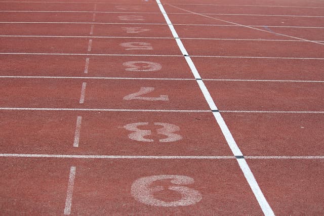 Close-up of a running tracks finish line with numbered lanes visible in white on the reddish-brown surface. The numbers are arranged from 3 to 8 in descending order from left to right. Lines run horizontally across the lanes.