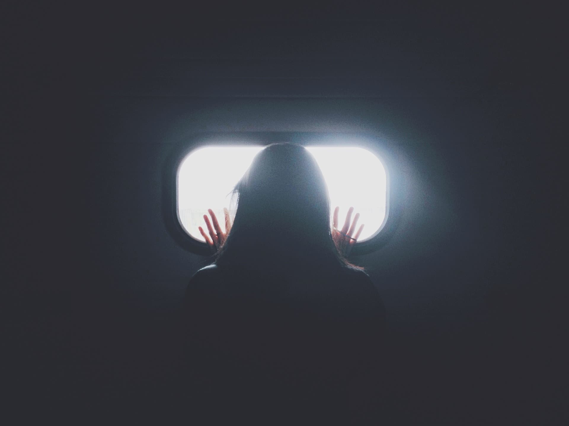 Silhouette of a person facing a bright window, with hands touching the glass. The dark room contrasts with the light streaming from the window, casting a soft glow around the figure.
