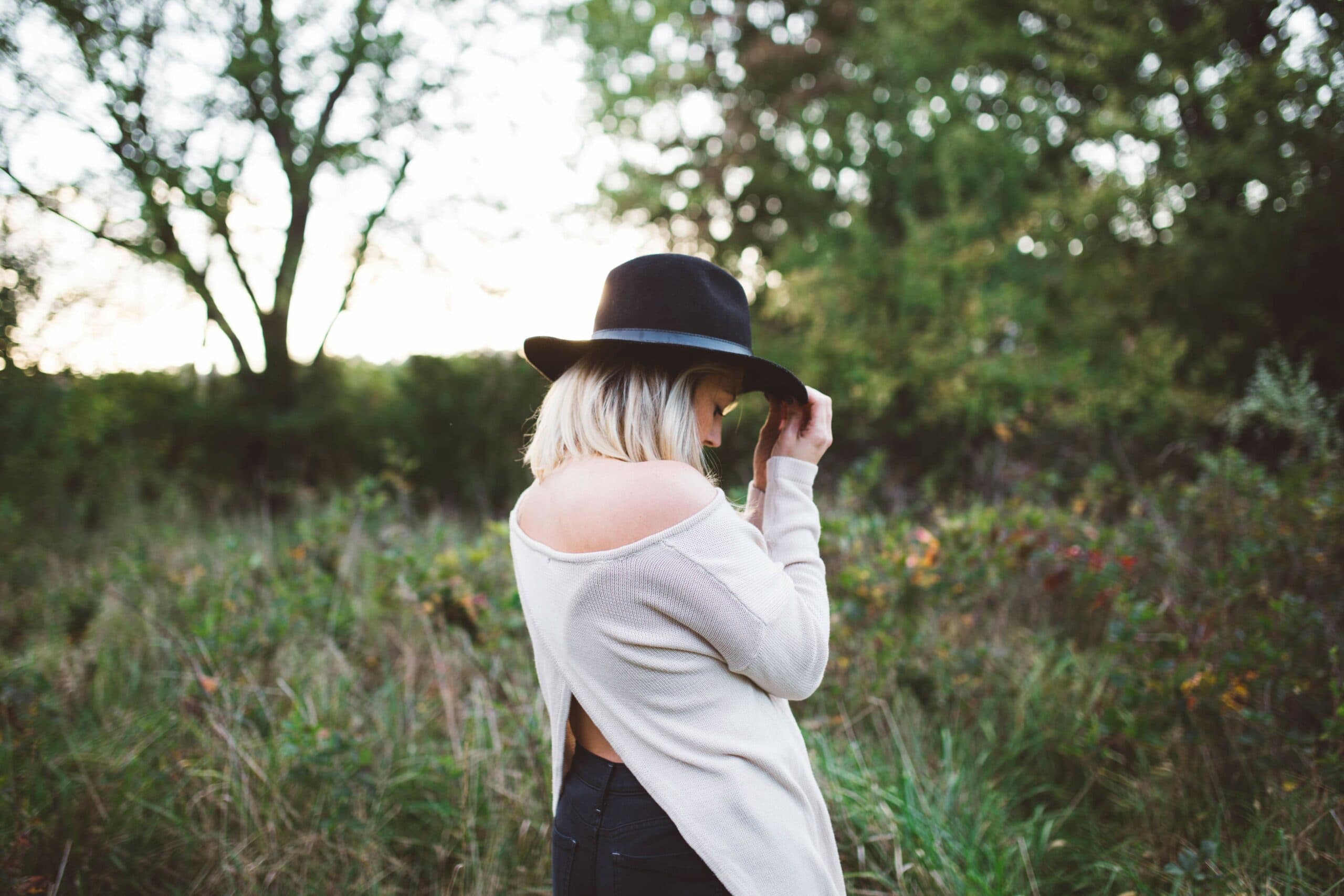 A person with blonde hair wearing a black hat and an off-shoulder sweater stands in a grassy field, surrounded by trees. The person is turned away, looking down, with a serene natural backdrop in soft daylight.
