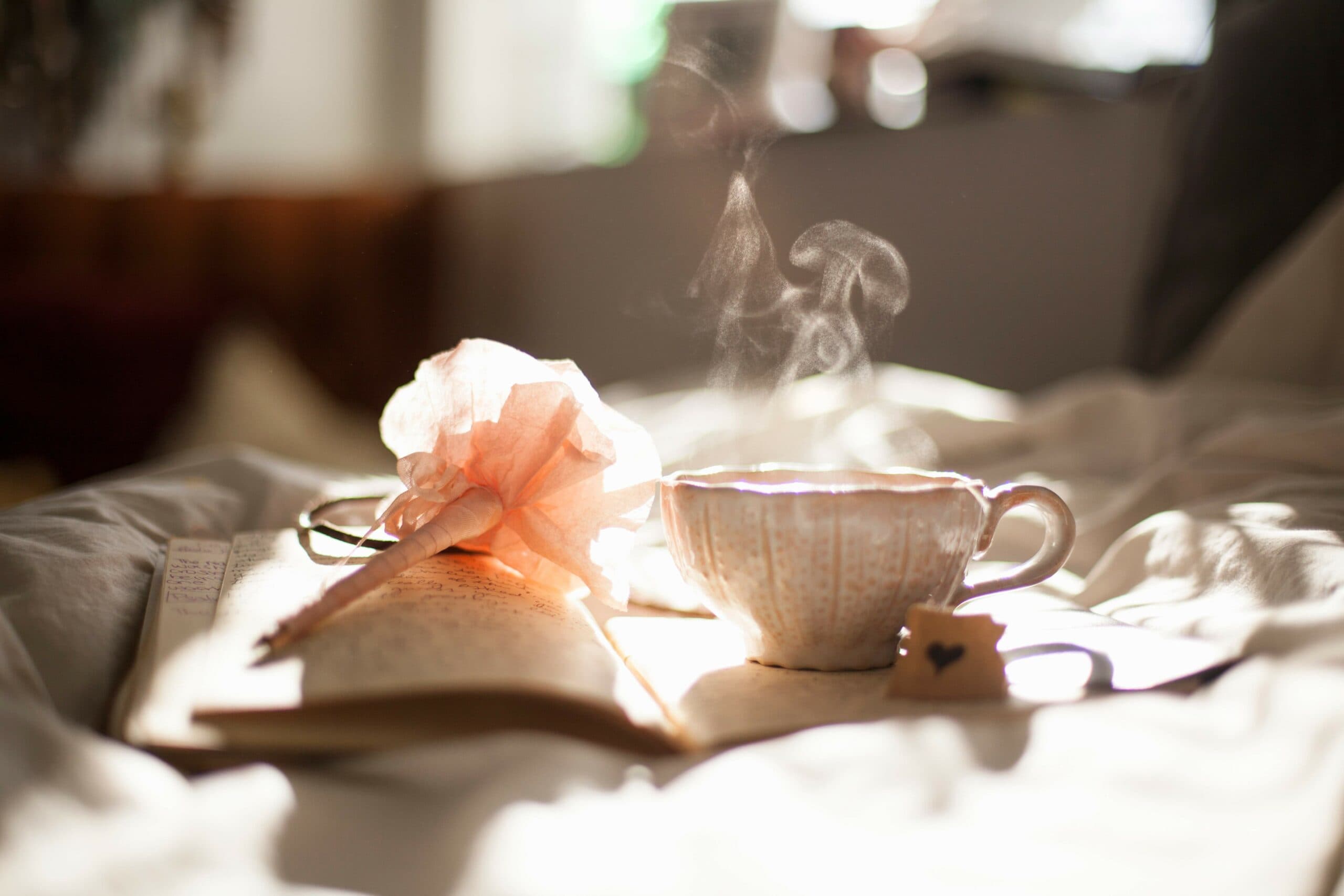 A steaming cup of tea sits on an open notebook, surrounded by soft light. A delicate pink flower rests nearby, creating a cozy and serene atmosphere.