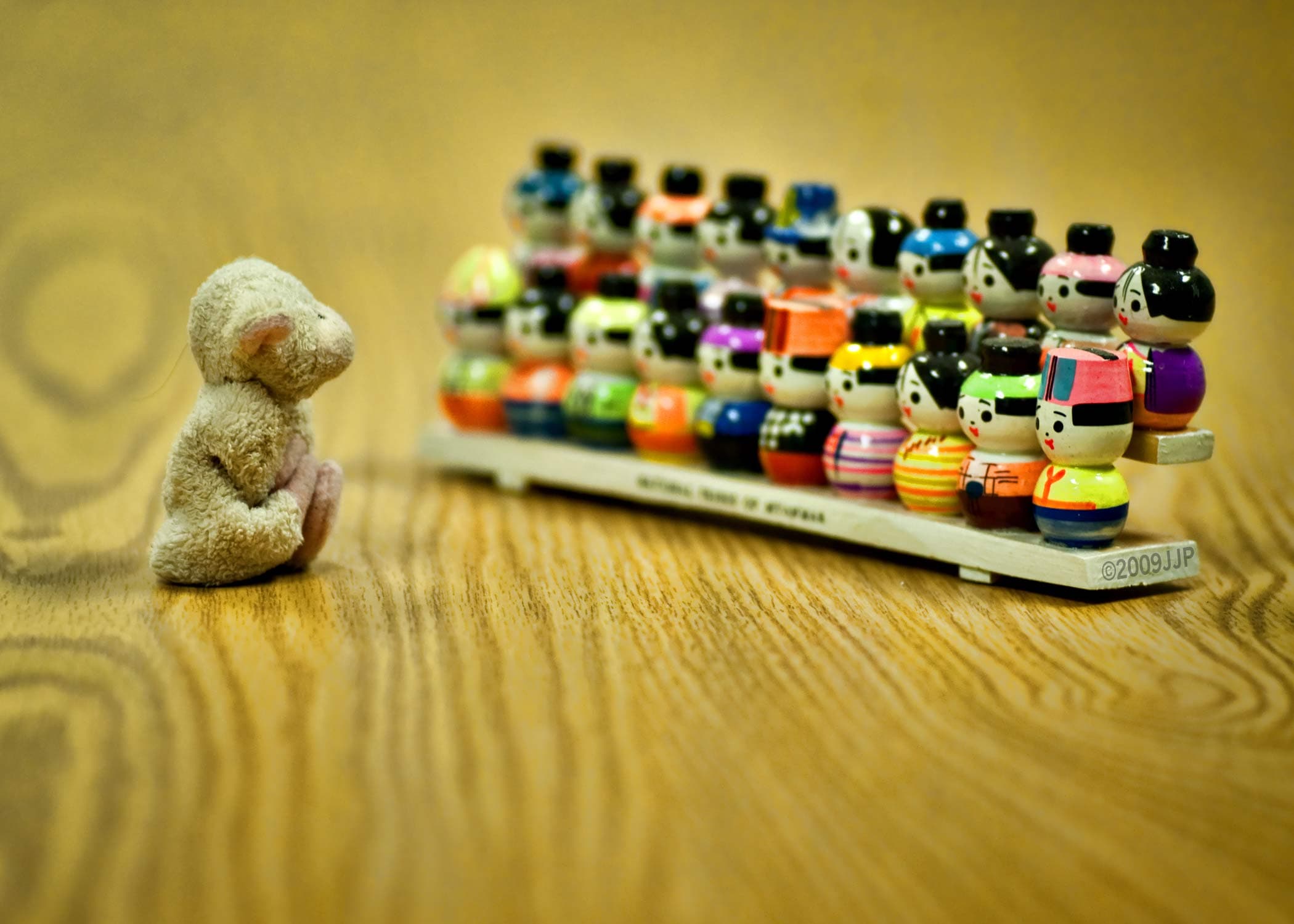 A small plush mouse sits on a wooden surface facing a row of colorful, miniature wooden dolls representing diverse figures. The dolls are arranged neatly on a long base, each painted with unique patterns and colors.