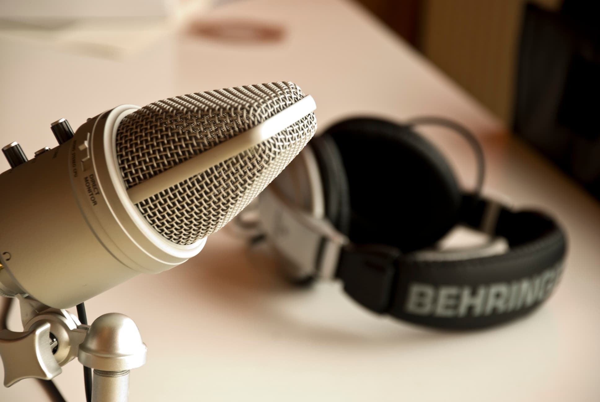 A close-up of a microphone with a metal mesh grill on a stand, positioned on a white surface. In the background, a pair of over-ear headphones with Behringer written on the headband is resting on the same surface.