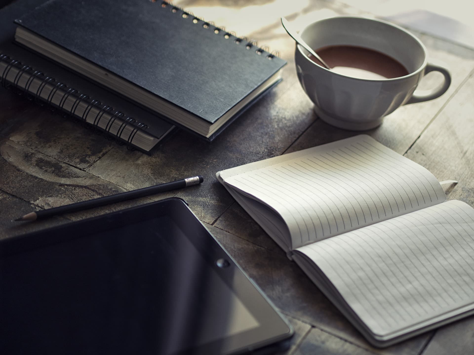 A serene workspace featuring a tablet, a closed spiral notebook, an open lined notebook with a pencil, and a cup of coffee on a rustic wooden table. Sunlight softly illuminates the scene, creating a calm and inviting atmosphere.