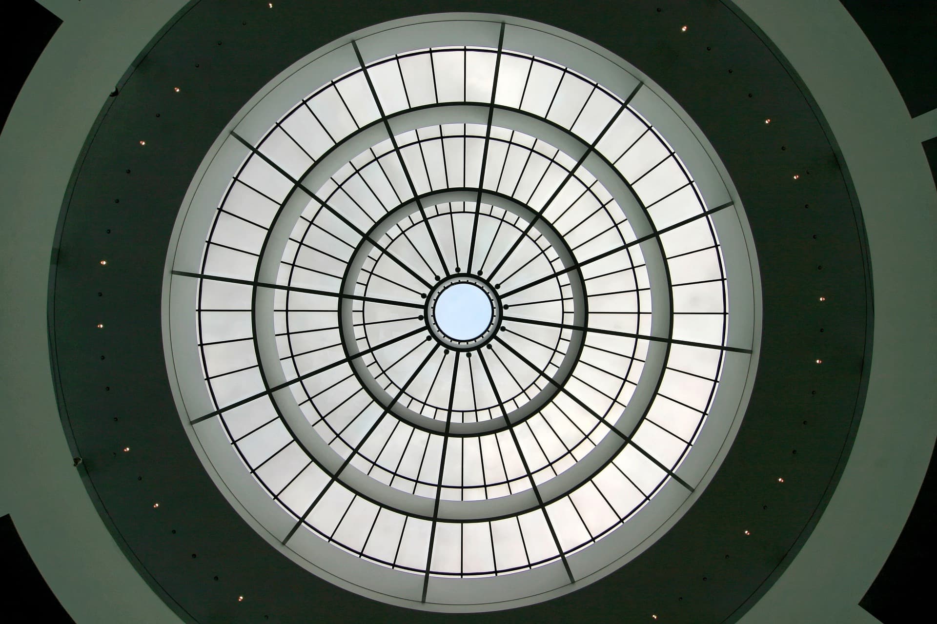 A view of a circular, geometric ceiling from below. It consists of concentric rings with radiating lines, creating a spiral effect. The central circle is blue-lit, while surrounding areas are illuminated by natural light through the glass panels.