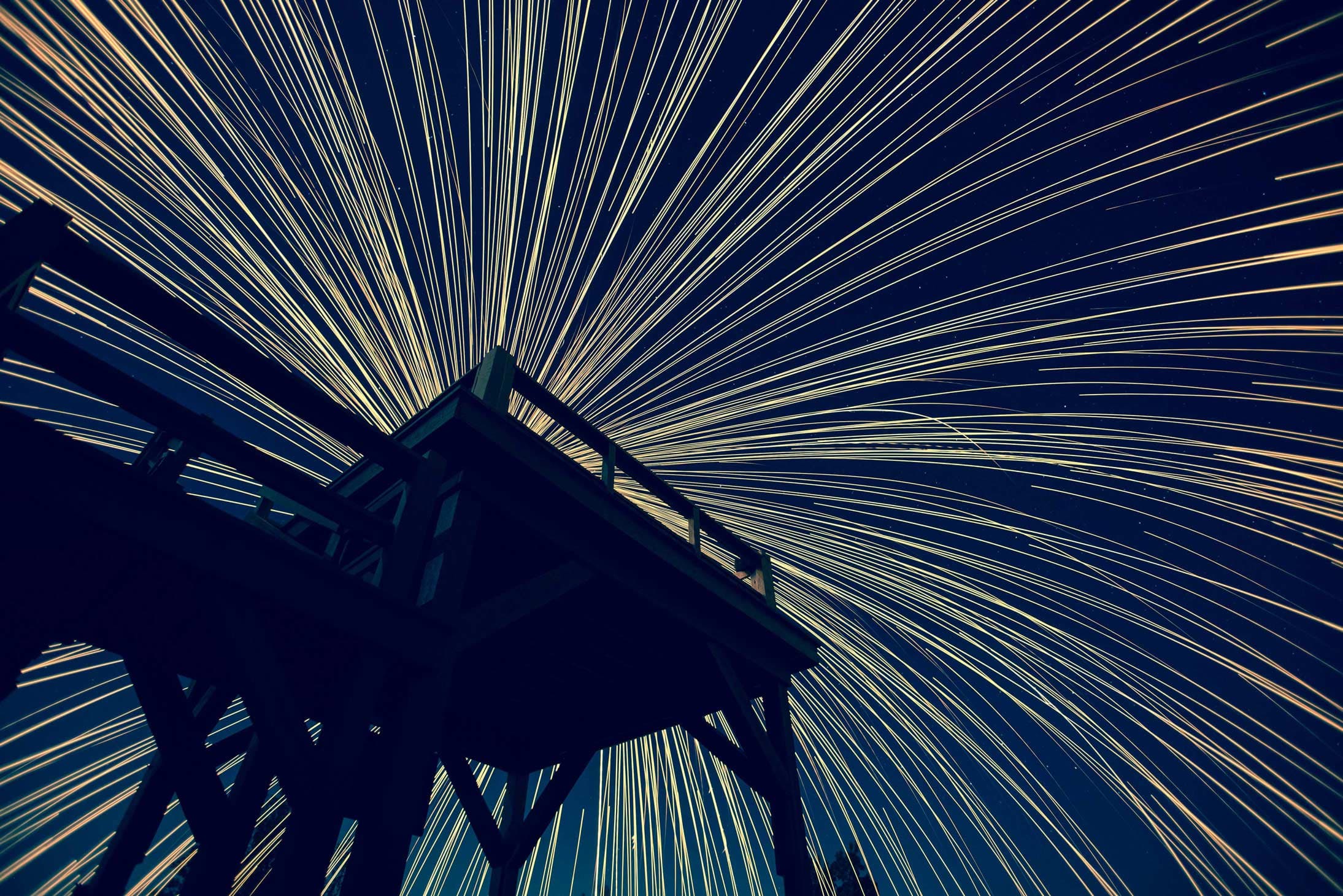 A wooden observation deck silhouetted against a night sky filled with striking star trails, creating a mesmerizing radial pattern. The long exposure captures the rotation of the Earth, making the stars streak across the image.