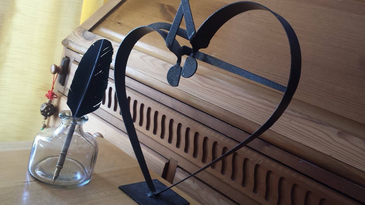 A black metal sculpture shaped like a heart with a keyhole sits on a wooden table. Beside it is a glass inkwell holding a quill. A wooden piece with carved details is in the background.