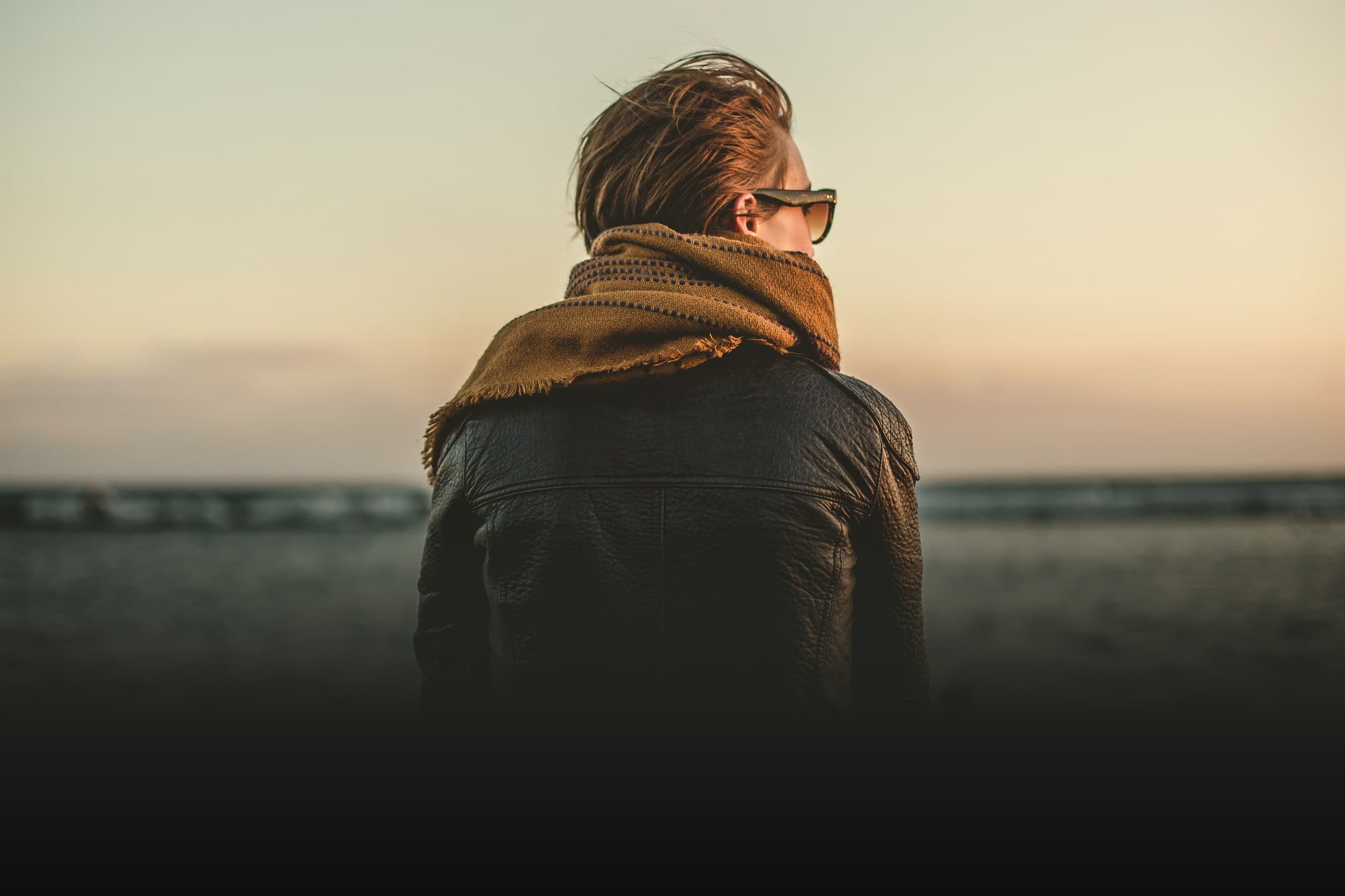 A person wearing sunglasses and a scarf is facing away, standing on a beach. The individual is dressed in a dark jacket, with their short hair tousled by the wind. The sky is hazy, with the sun setting in the background.