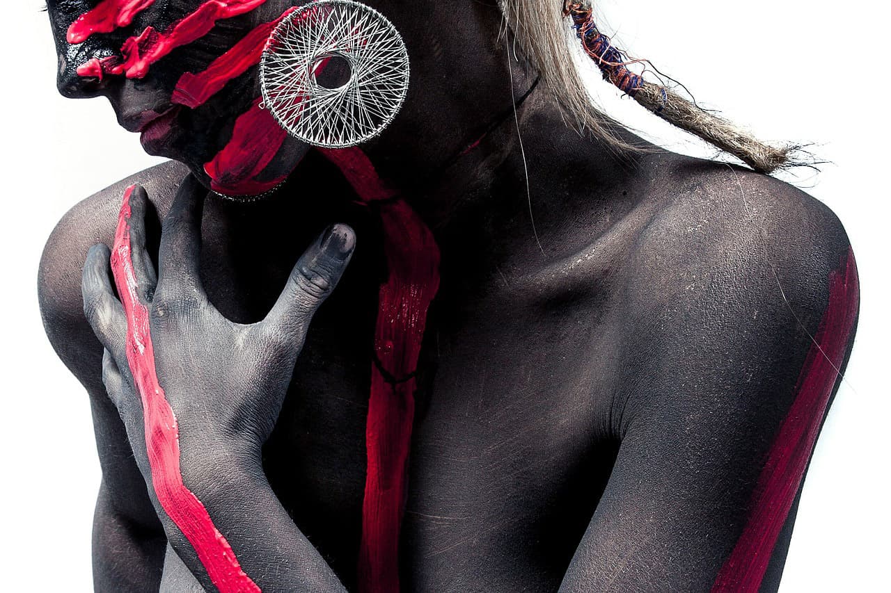 A person with body paint, predominantly black with red stripes across the face and body, wears a large circular earring. The hair is partially braided, and the pose is contemplative, with one hand on the chest against a white background.