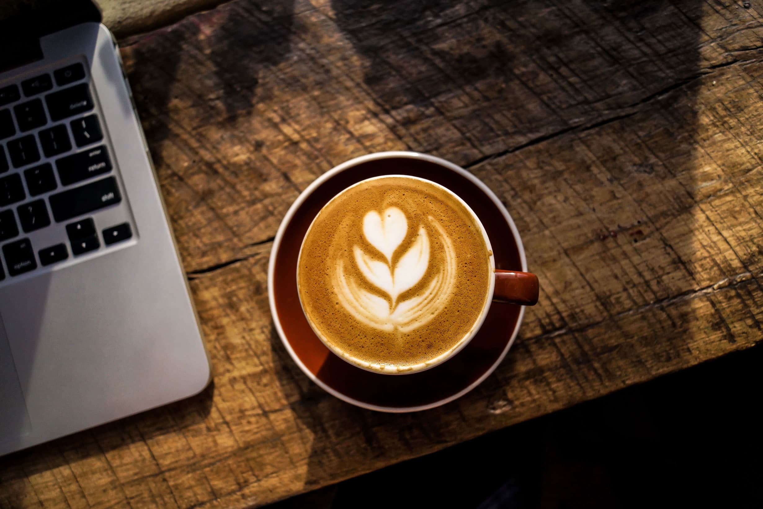 A cup of latte with a heart and leaf design in the foam sits on a wooden table. A corner of a laptop is visible to the left, with soft lighting casting shadows on the table.