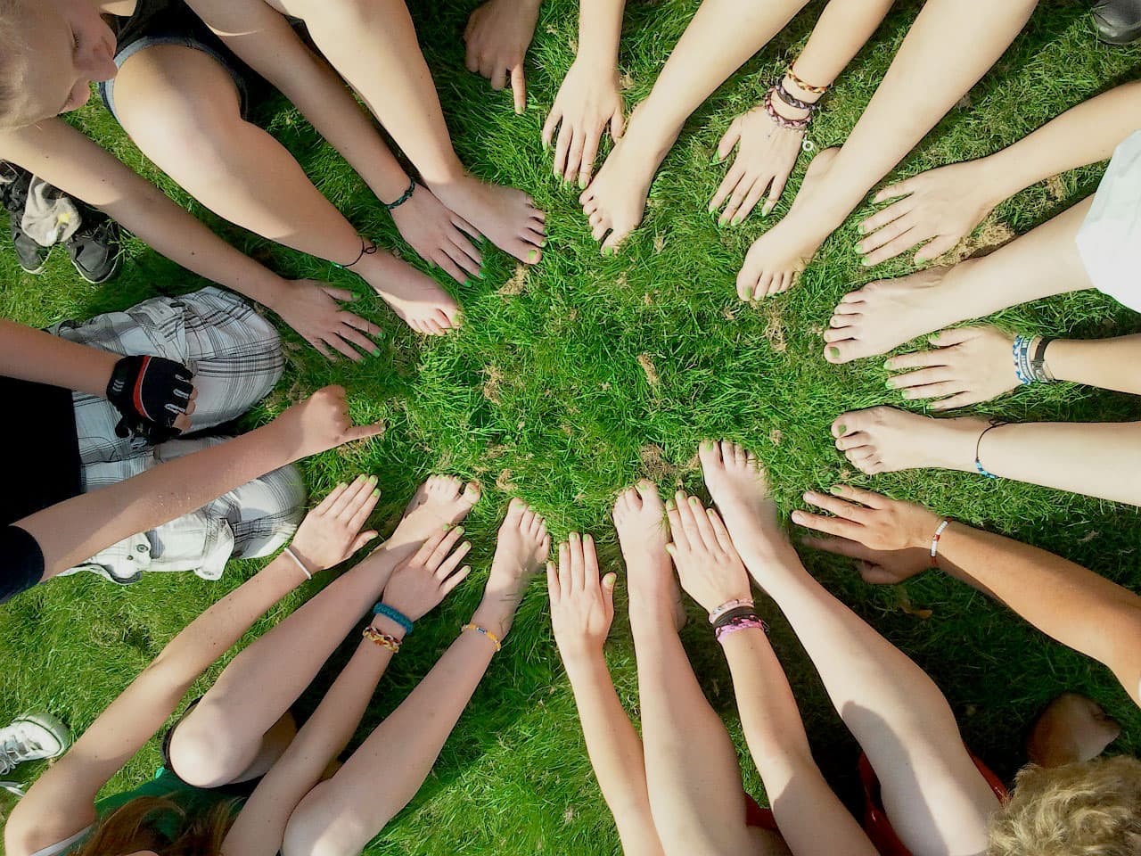 A group of people sit in a circle on grass, extending their hands and bare feet toward the center. The scene conveys unity and togetherness, with various bracelets on some wrists.
