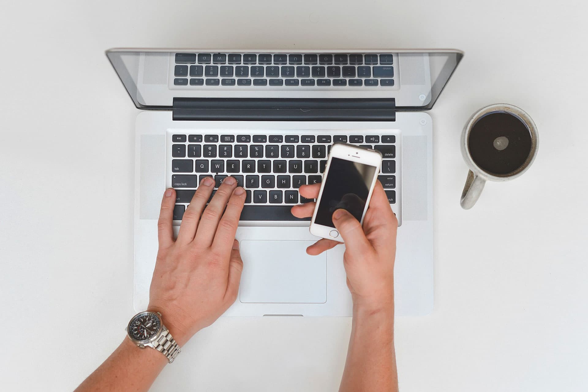 Two hands are seen working on a laptop keyboard while holding a smartphone. A cup of coffee sits nearby on a white surface. The person is wearing a watch on their left wrist. The setting suggests a workspace.