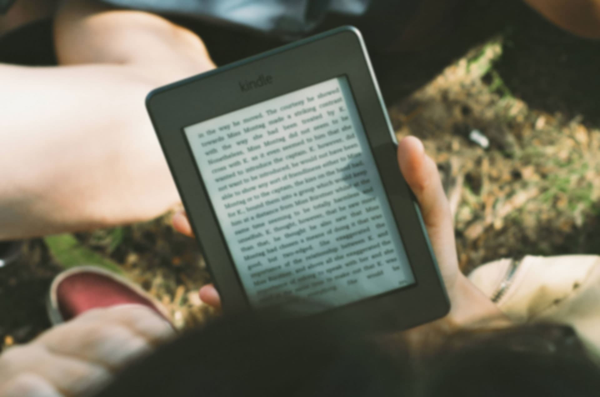 Person holding a Kindle e-reader displaying text, sitting outdoors on grass, with legs and part of another person visible in the background. The scene suggests reading in a relaxed, natural setting.