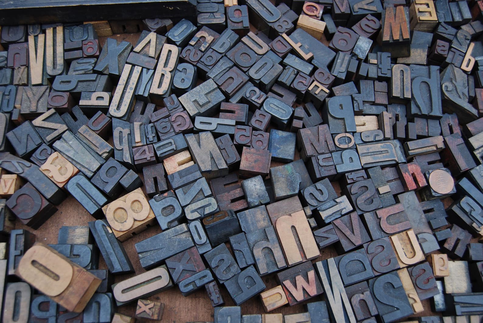 A scattered pile of vintage wooden letterpress blocks in various sizes and fonts, displaying letters and numbers in different orientations and shades of brown and black. The image is rich in texture and complexity.