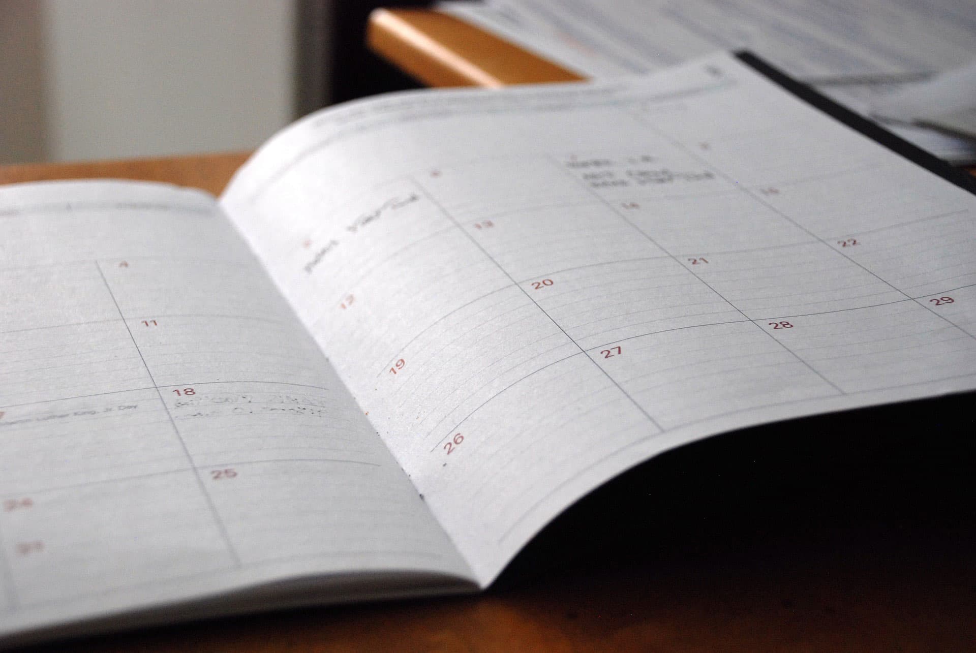 An open planner lies on a wooden surface, displaying a monthly calendar layout. The pages are blank, with dates visible and some faint handwritten notes in the margins. The background is softly blurred.