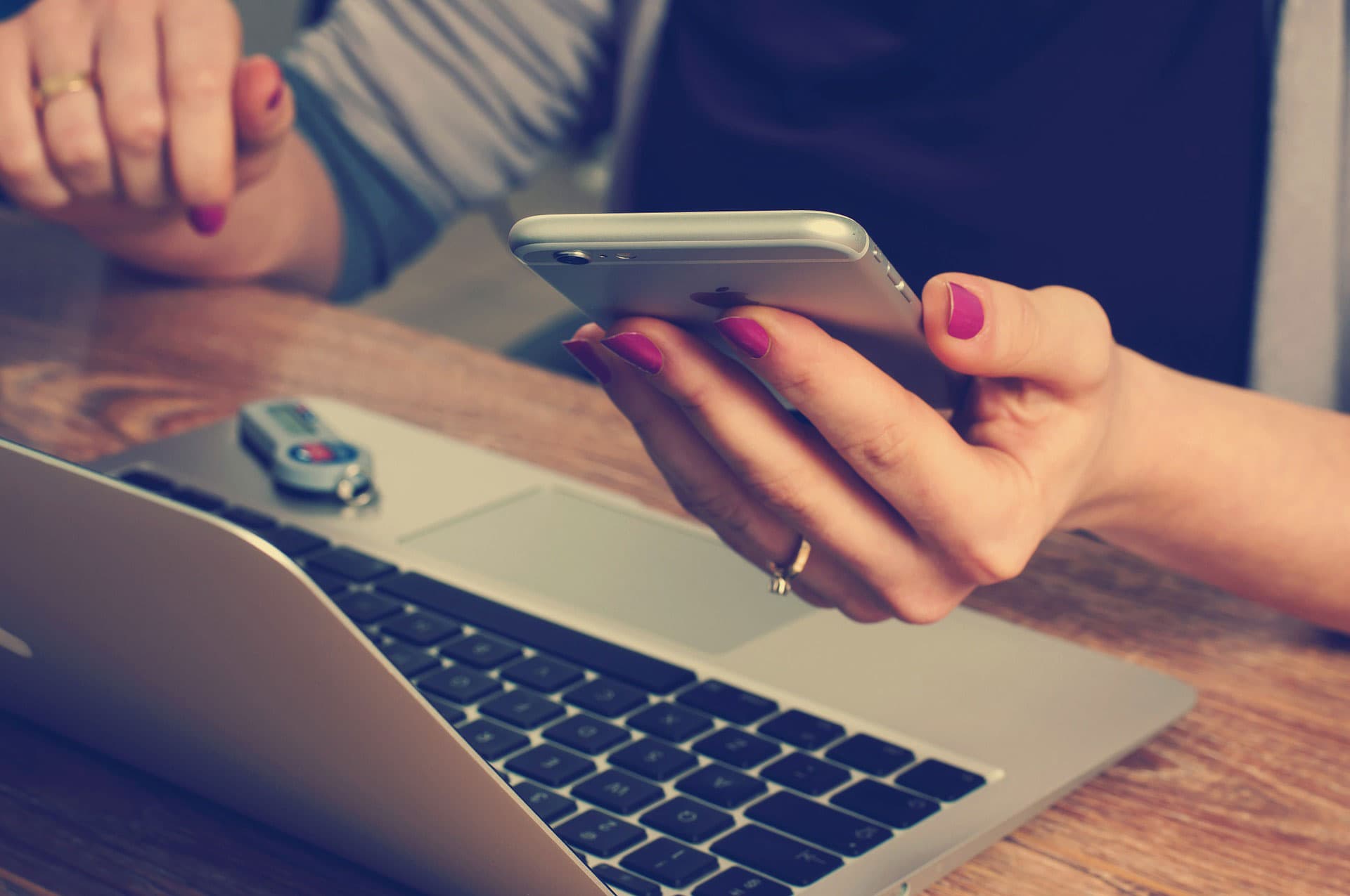 A close-up of a person holding a smartphone in one hand while using a laptop, with a USB flash drive plugged into the laptop. The person has manicured nails and wears a ring. The setting suggests a workspace or office environment.