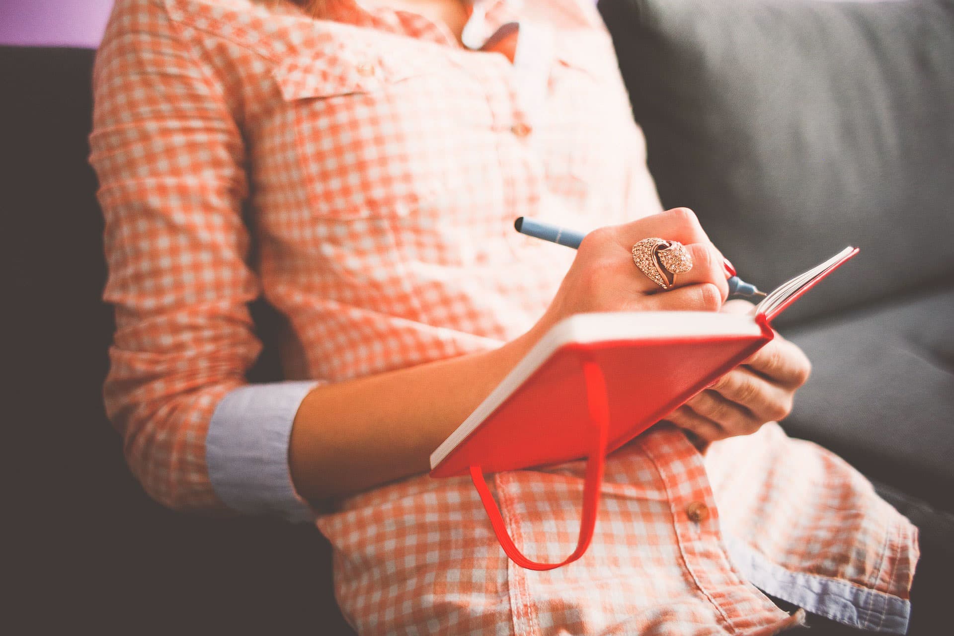 A person sitting on a couch writes in an open red notebook with a blue pen. They are wearing a peach-colored plaid shirt and a heart-shaped ring. The setting appears relaxed and informal.
