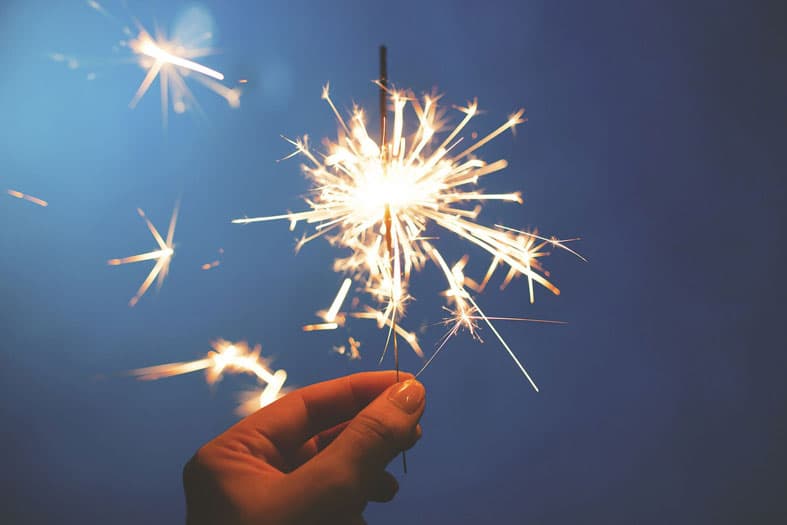 A hand holding a glowing sparkler against a dark blue background, with bright sparks radiating outward.