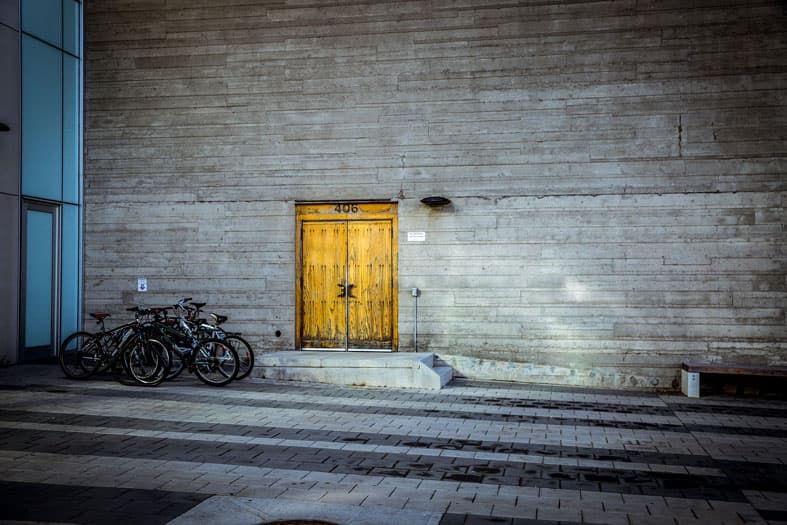 A concrete wall with a yellow wooden door labeled 406 at the center. Four bicycles are parked to the left of the door on a paved area, with dim lighting casting shadows on the wall.
