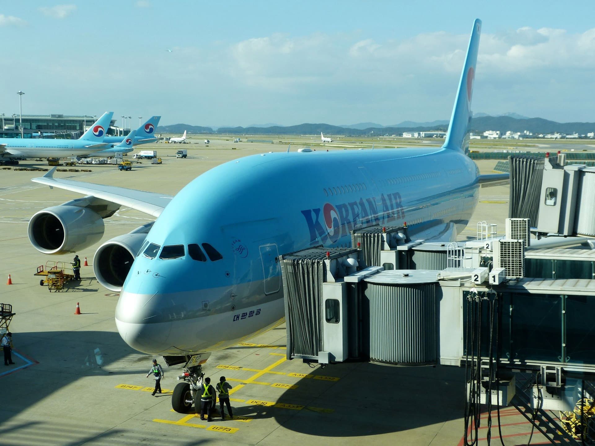 A Korean Air airplane is parked at an airport gate. Ground crew members are near the planes nose. The sky is clear, and another airplane is visible in the background on the runway.