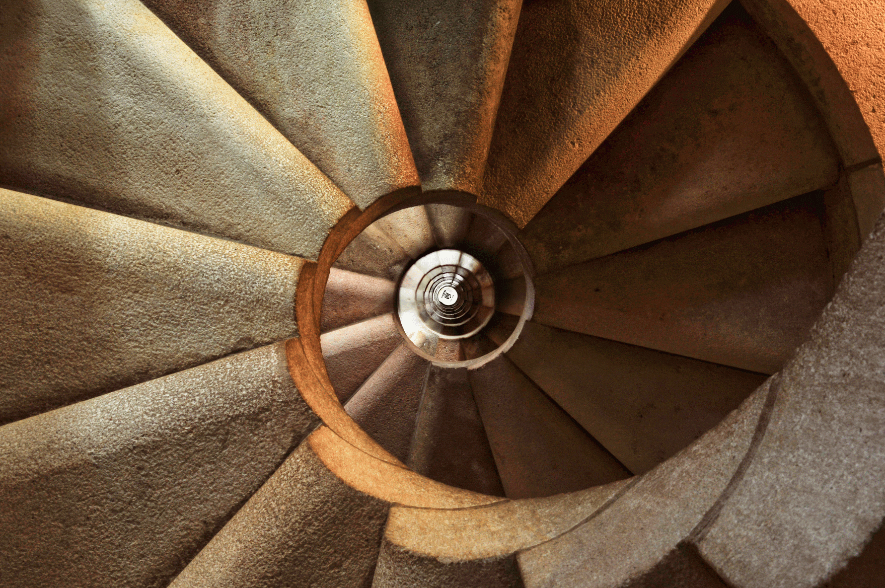 A spiraling staircase viewed from above, showcasing a circular pattern with warm stone colors. The spiral creates a captivating geometric design, drawing the eye towards the central point at the bottom.