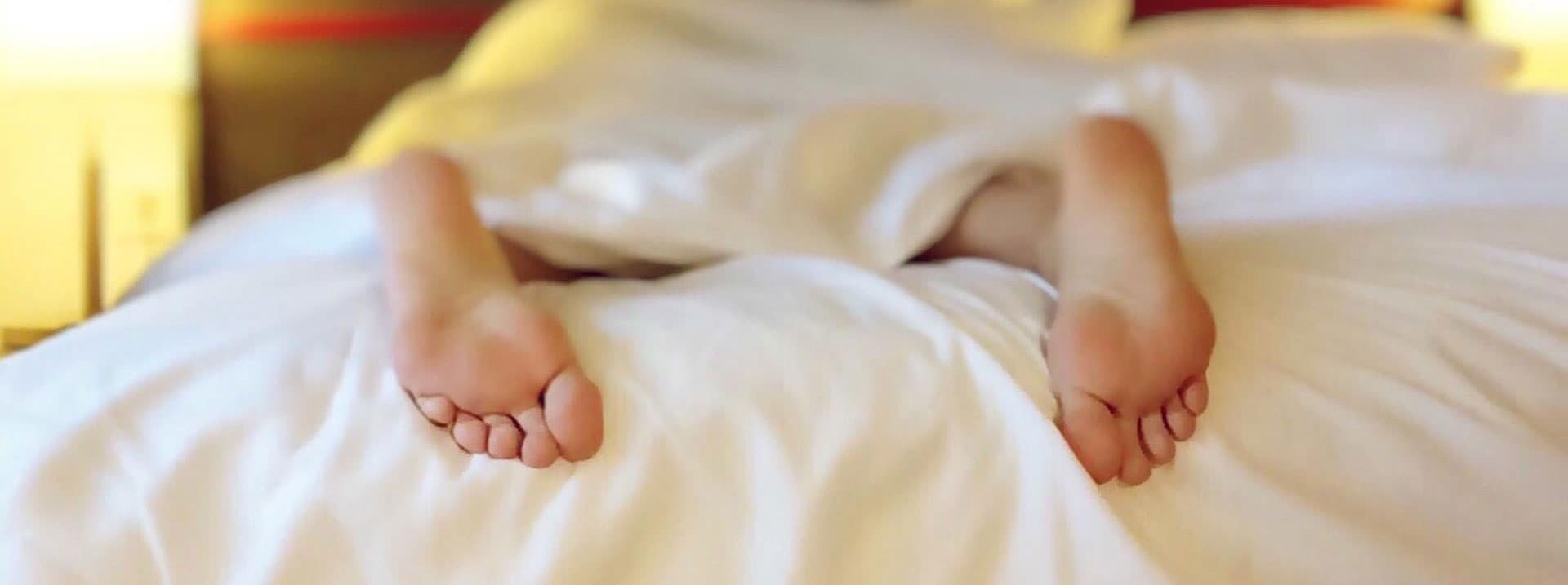 Person lying in bed with bare feet sticking out from under a white blanket, in a softly lit room.
