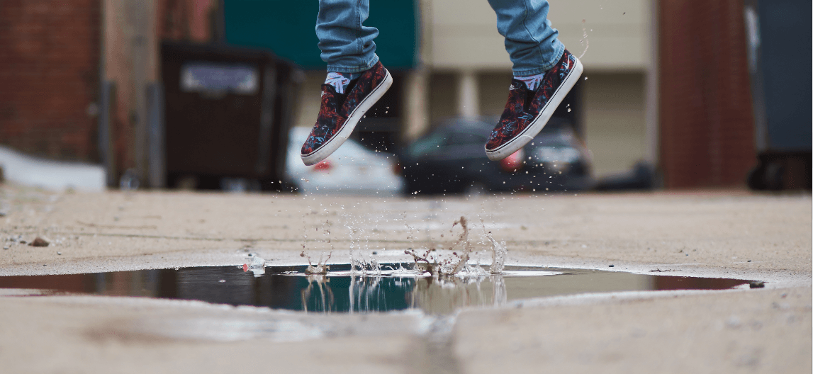 Person wearing jeans and patterned sneakers jumping over a puddle on a concrete path, causing splashes of water. Urban background with blurred buildings.