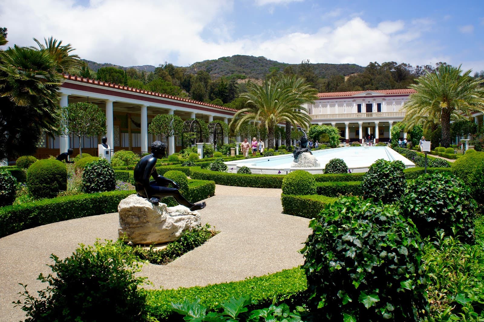 Luxurious garden outside a classical building with columns, featuring neatly trimmed hedges, palm trees, and a central pool. A dark statue sits on a rock near the pool, with mountains and blue sky in the background.