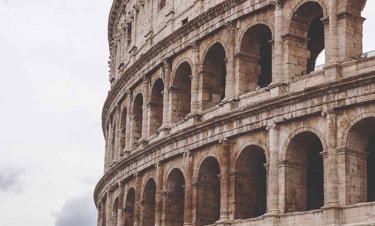 Partial view of the Colosseum in Rome, Italy, showcasing its ancient stone arches and weathered exterior. The sky is overcast, highlighting the iconic architecture against the muted background.
