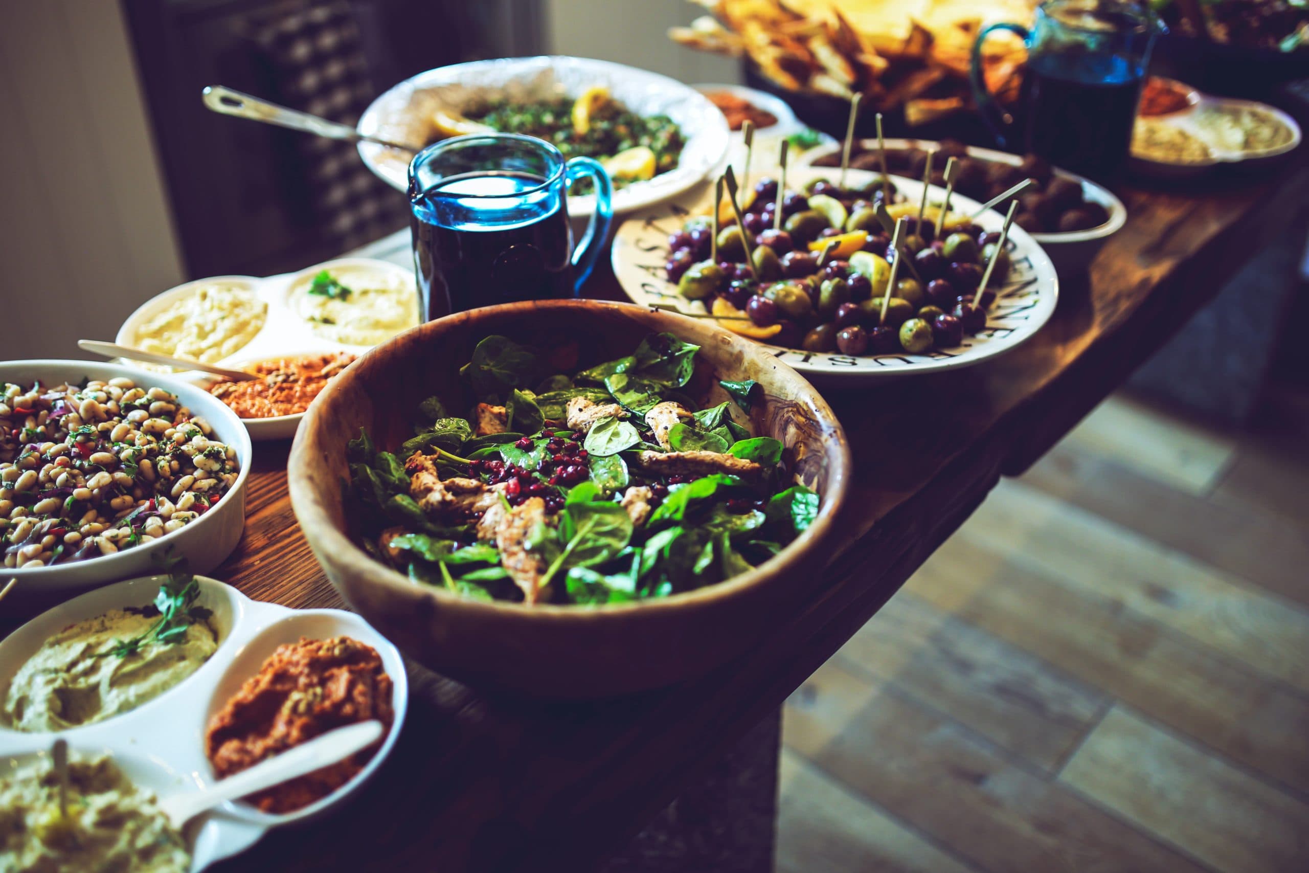 A wooden table filled with various dishes: a large salad bowl, plates of stuffed olives, small bowls of dips, and other assorted foods. The table is set indoors with soft lighting.