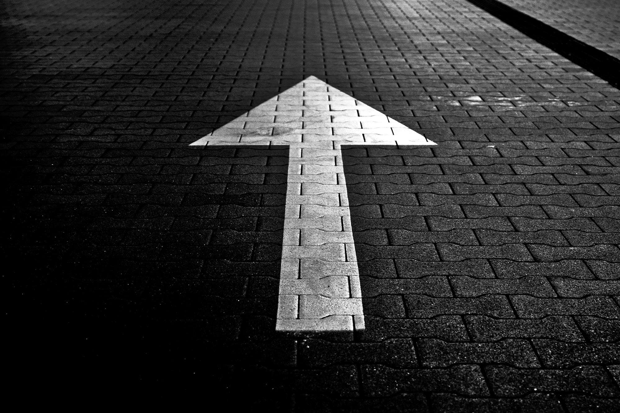 A textured brick pavement with a large white arrow painted on it, pointing upwards. The image is in black and white, creating a high-contrast effect between the arrow and the surface.