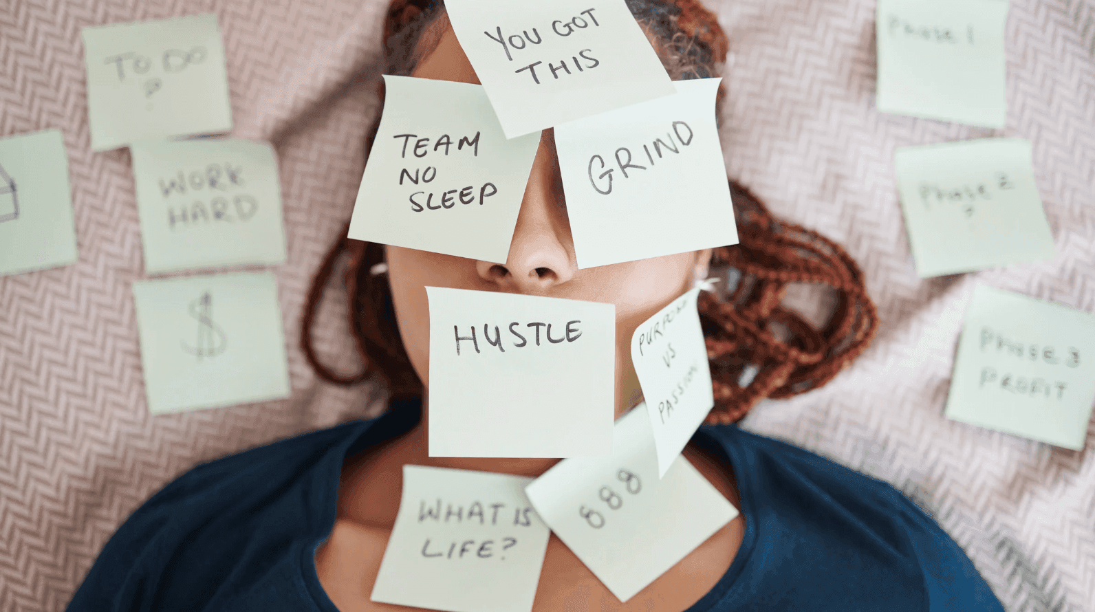 A person lies on a surface with sticky notes covering their face, displaying phrases like You got this, Hustle, and Team no sleep. Additional notes surround them with words such as Grind and Work hard.