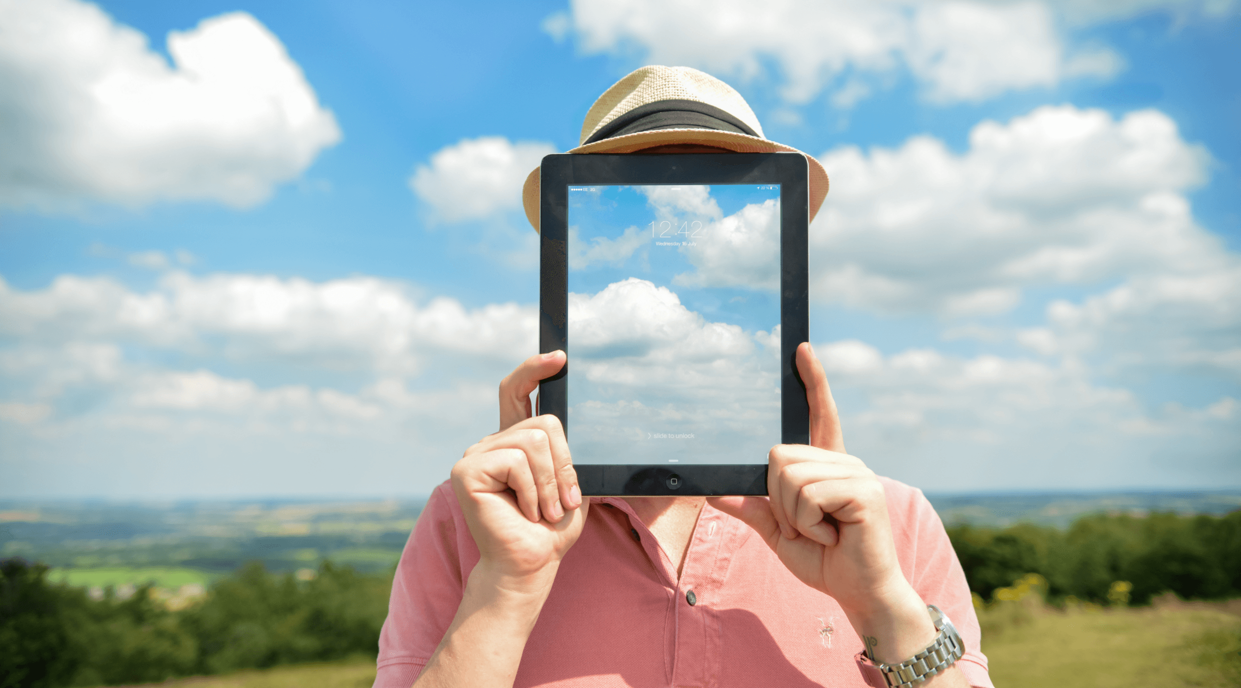 A person wearing a hat holds a tablet in front of their face. The tablet screen reflects a blue sky with fluffy clouds, blending with the actual sky in the background. The person is standing outdoors on a clear day.