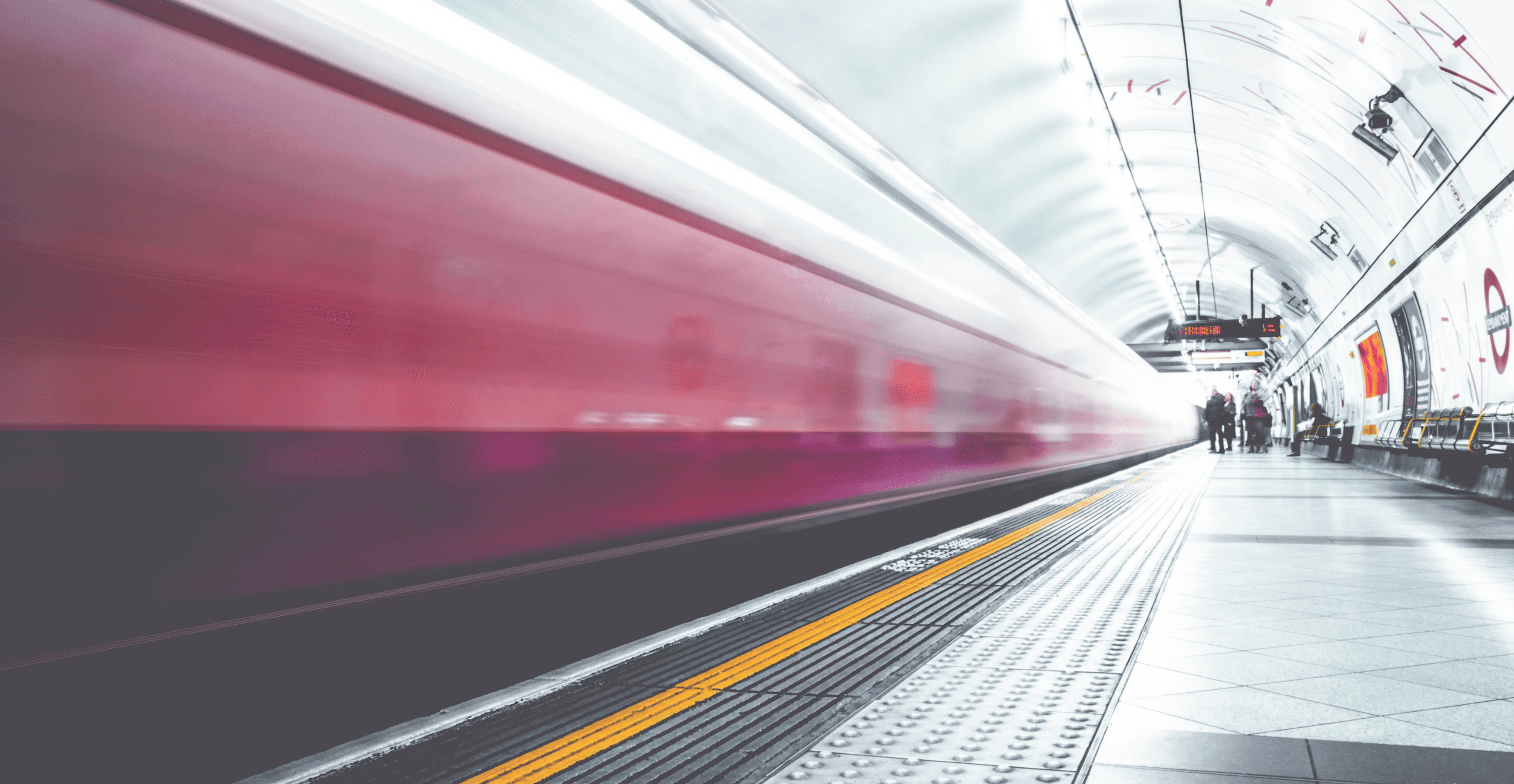A subway train speeds through a brightly lit underground station, creating a motion blur effect. In the background, a few people stand on the platform. The scene is vibrant, with red and white colors dominating the stations design.