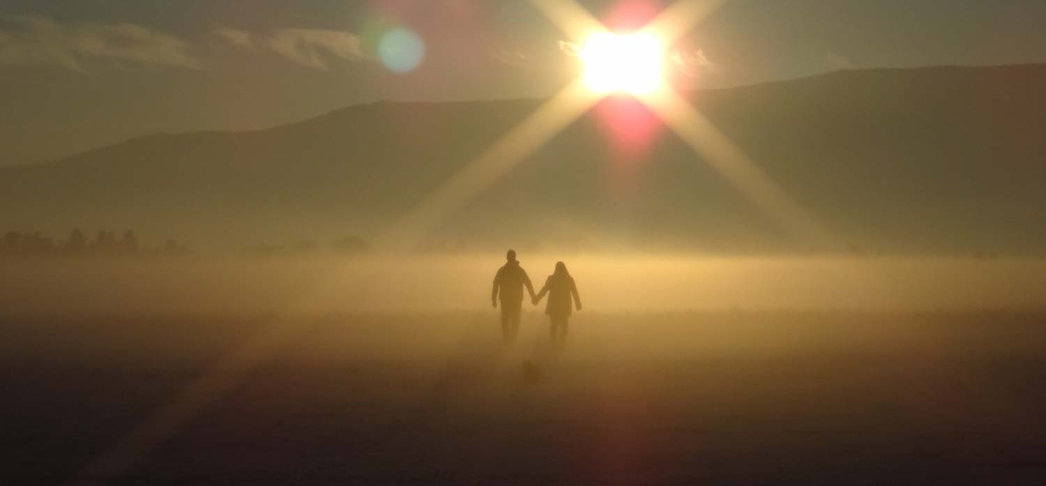 A couple walks hand in hand through a misty landscape at sunrise. The sun casts a bright glare, creating a golden glow around them and projecting long shadows on the ground. A mountain silhouette is visible in the background.