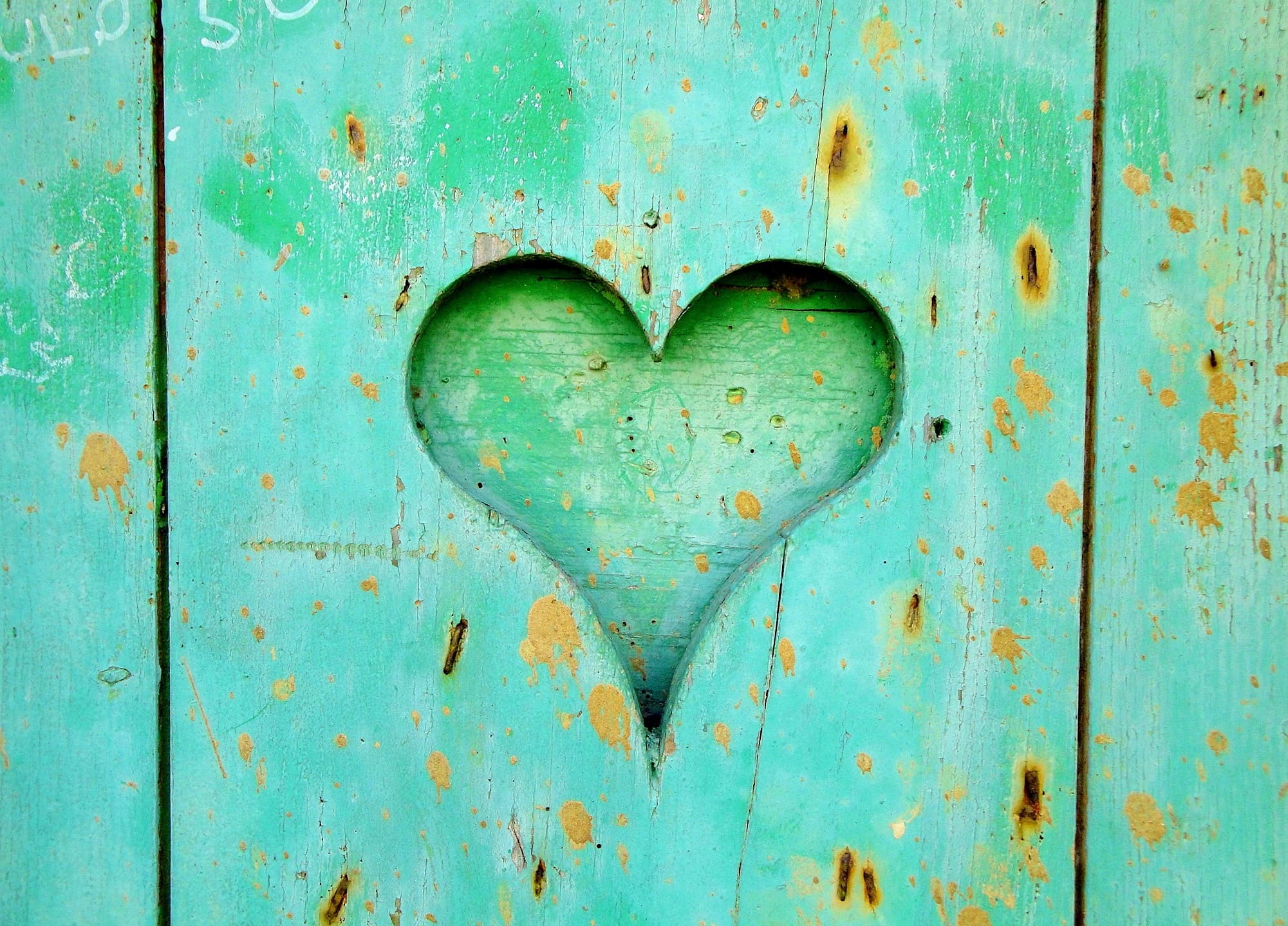 A heart-shaped cutout in a weathered, turquoise wooden surface, with rust-colored spots and visible grain patterns adding a rustic charm.