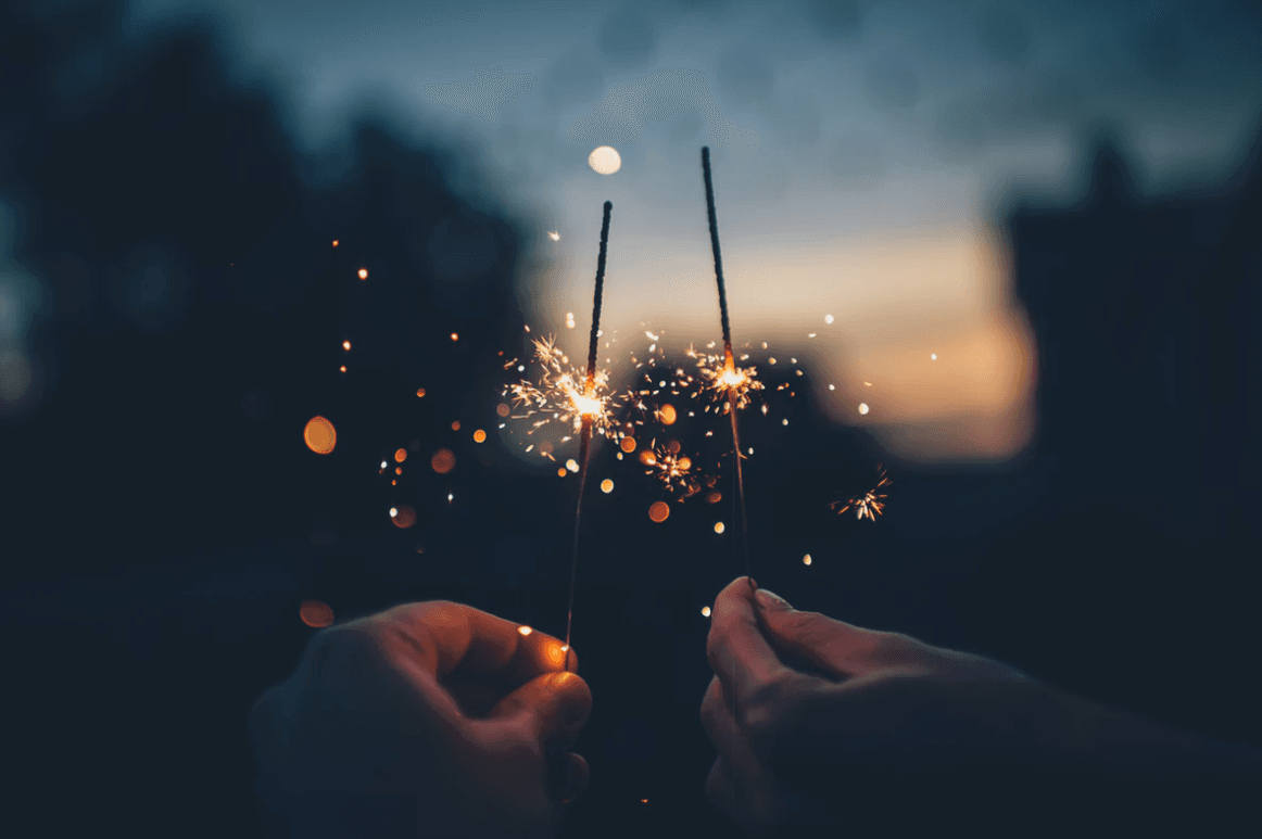 Two hands holding sparklers in a dark, outdoor setting with blurred lights in the background, creating a festive and warm atmosphere.