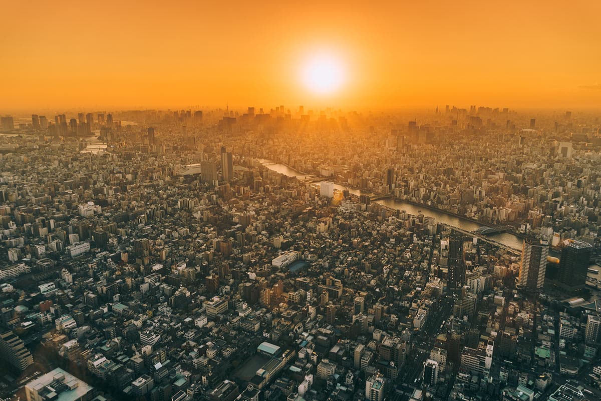 Aerial view of a sprawling cityscape at sunset with a river winding through the densely packed buildings. The sky is filled with orange hues as the sun sets on the horizon.