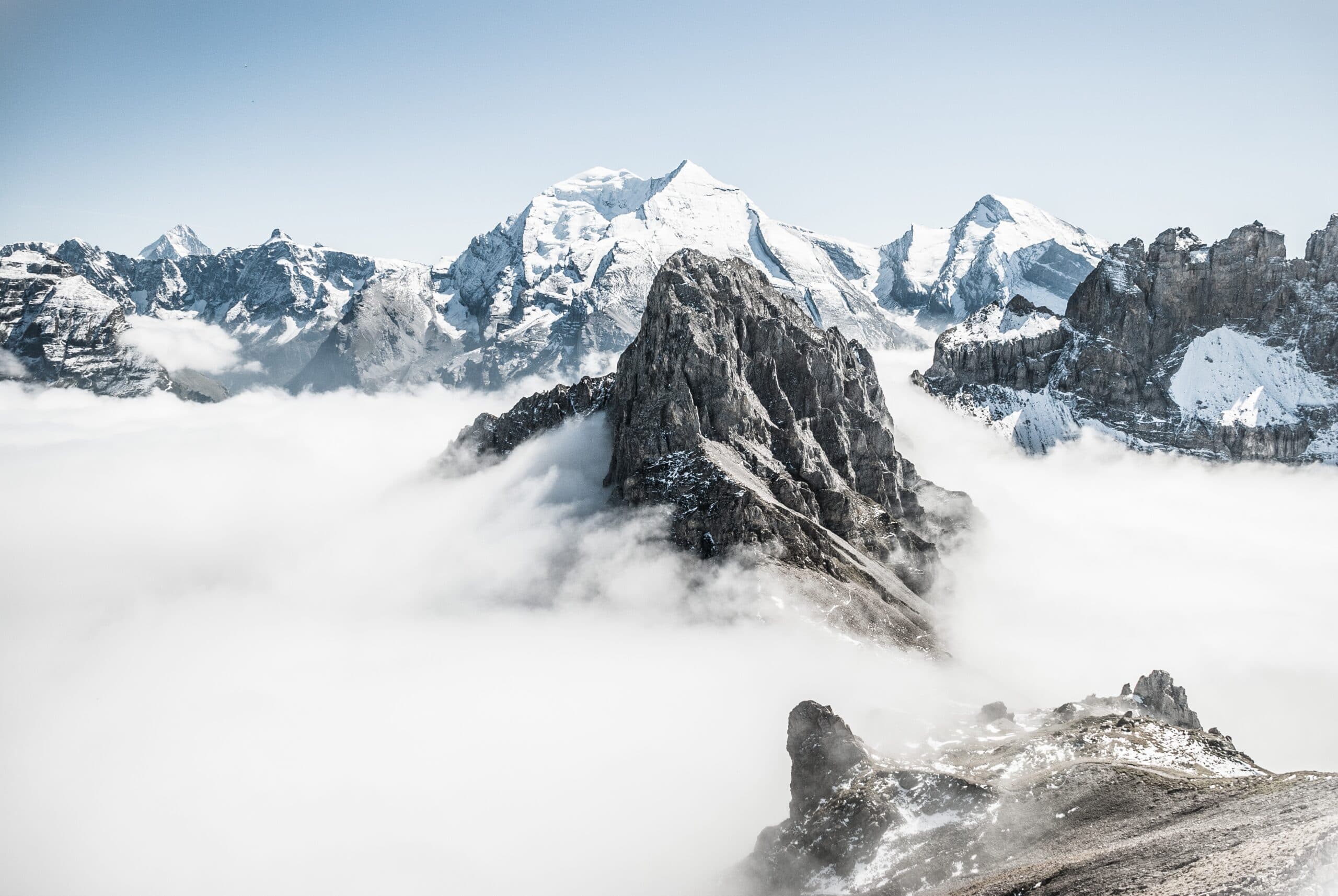 Snow-covered mountain peaks rise above a dense layer of clouds, with a prominent rocky summit in the foreground. The clear sky enhances the dramatic, serene landscape.