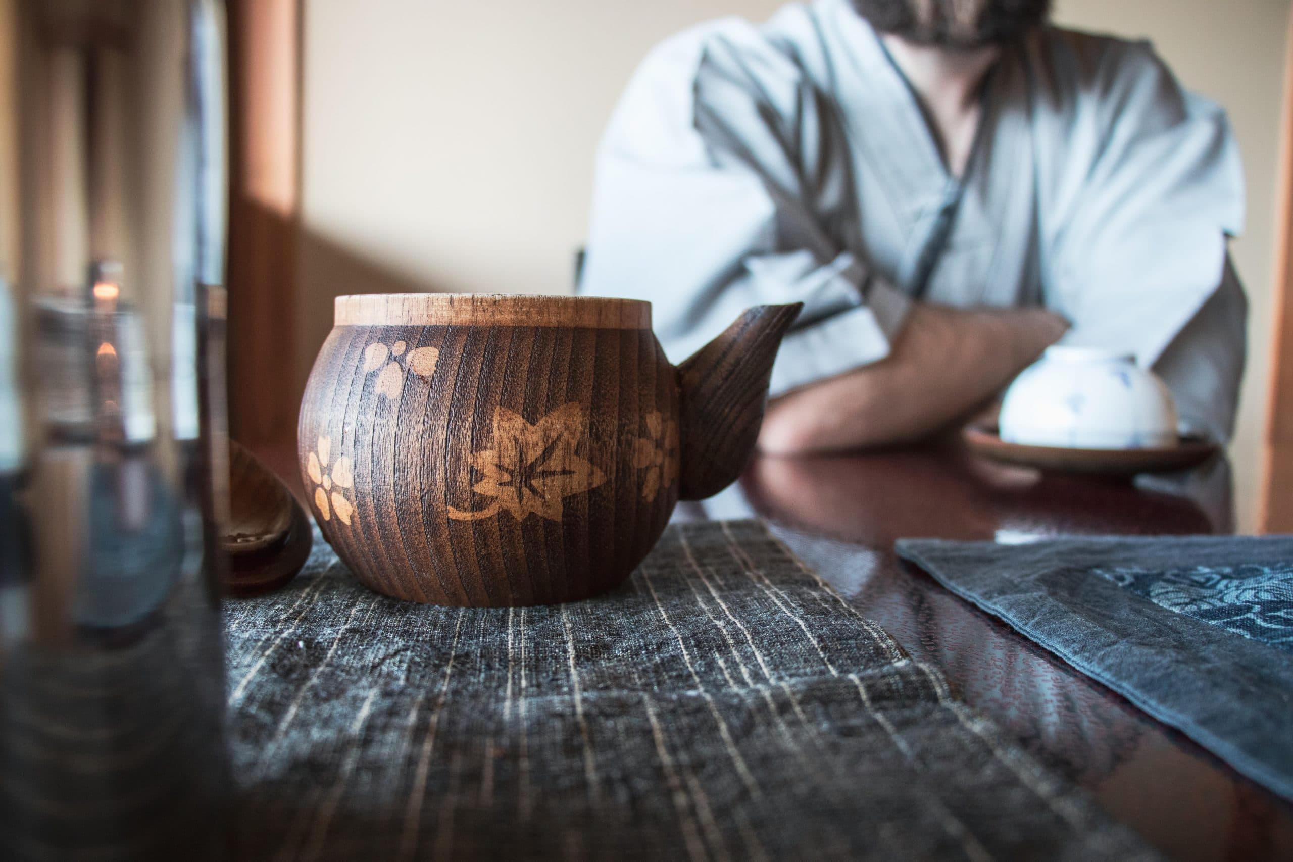 A wooden teapot with floral engravings sits on a table with woven placemats. In the background, a person in traditional clothing with folded arms is partially visible, holding a teacup. The scene conveys a tranquil, traditional setting.