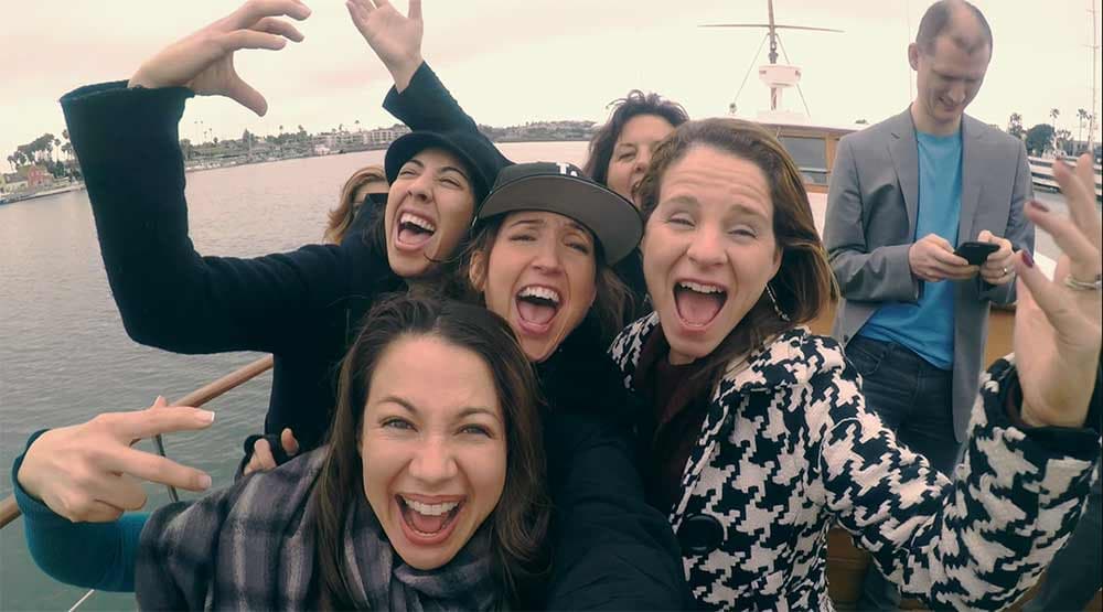 A group of five women and one man on a boat are joyfully posing for a selfie. The women are smiling widely and making playful gestures, while the man is focused on his phone in the background. The ocean and shoreline are visible behind them.