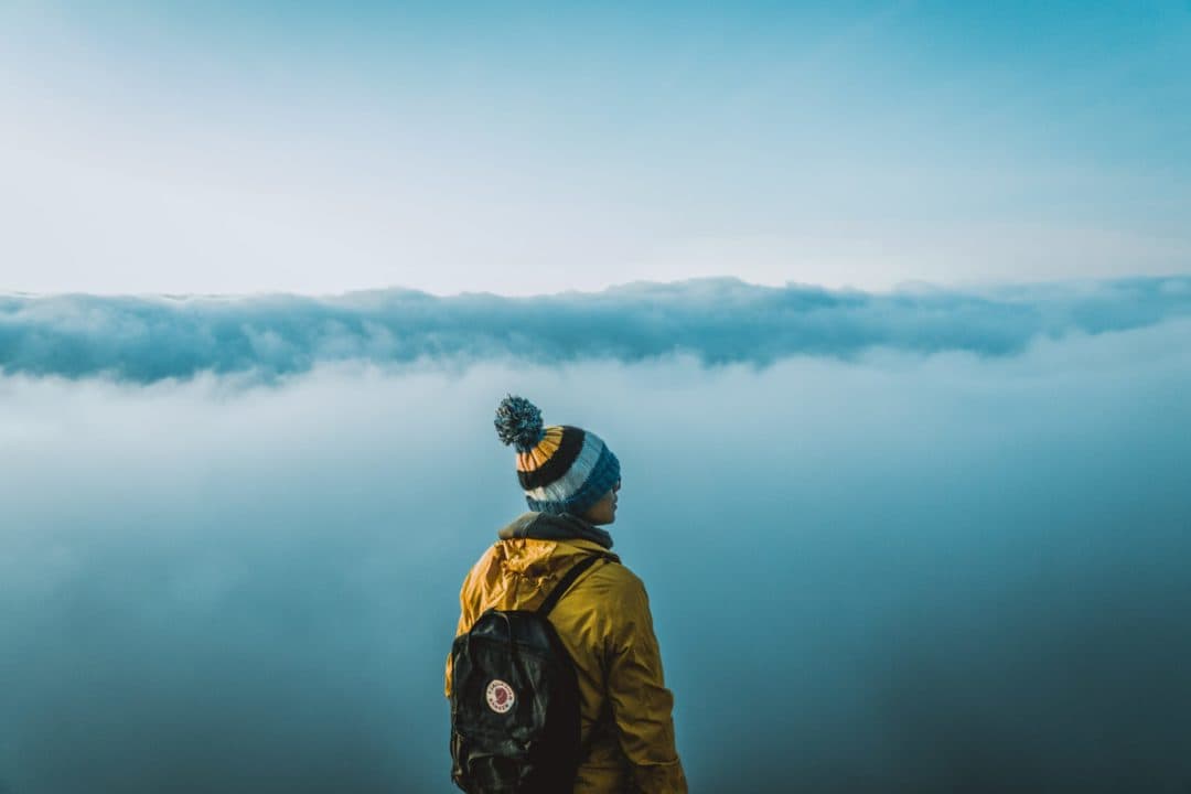 A person in a yellow jacket and pom-pom beanie stands on a cloud-covered mountaintop, gazing at the expansive blue sky above. A backpack is slung over their shoulder. The scene conveys a sense of exploration and tranquility.