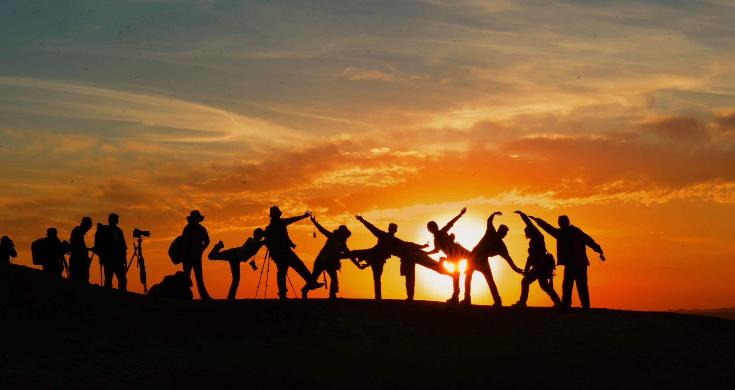 A group of people silhouetted against a vibrant sunset sky, standing on a hill. Some are holding cameras while others strike playful poses, creating a lively and dramatic scene.