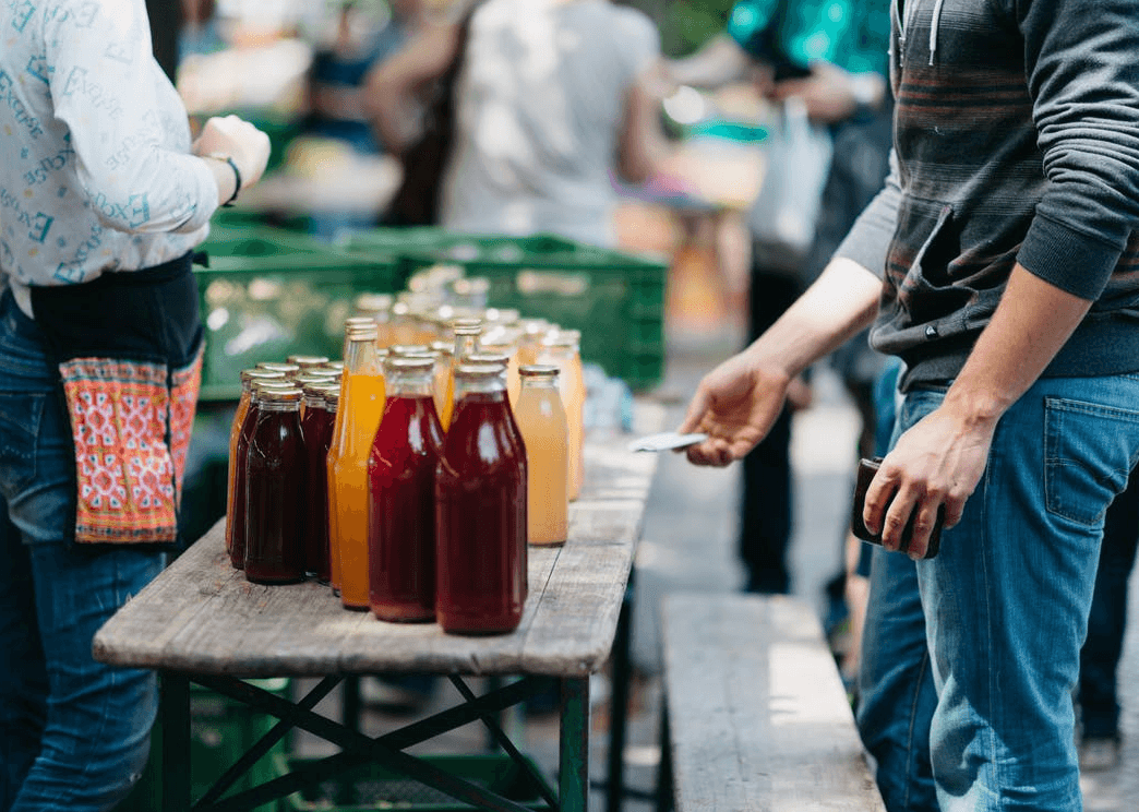 Customers at an outdoor market table purchase various bottled juices, including red, yellow, and orange varieties. One person holds a wallet, while another stands nearby. Blurred background shows more people and greenery.