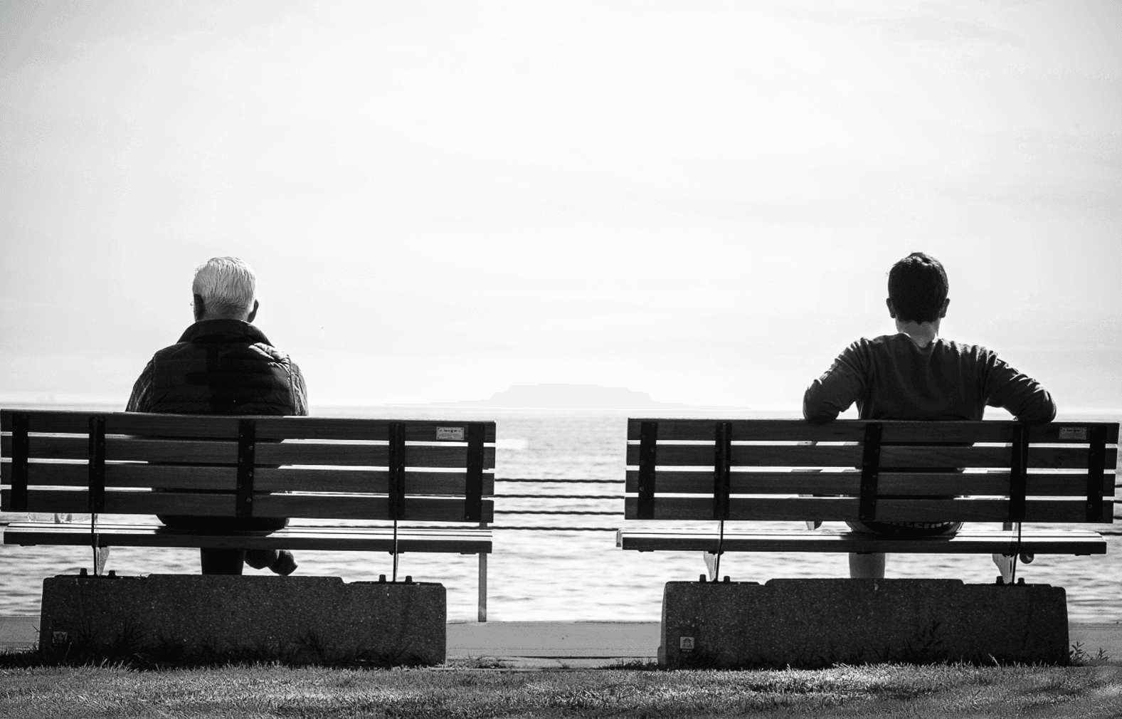 Two people sitting on separate benches, facing a calm sea. The person on the left has light-colored hair, and the person on the right has dark hair. The setting is peaceful and serene, with a clear horizon line under an open sky. Black and white photo.